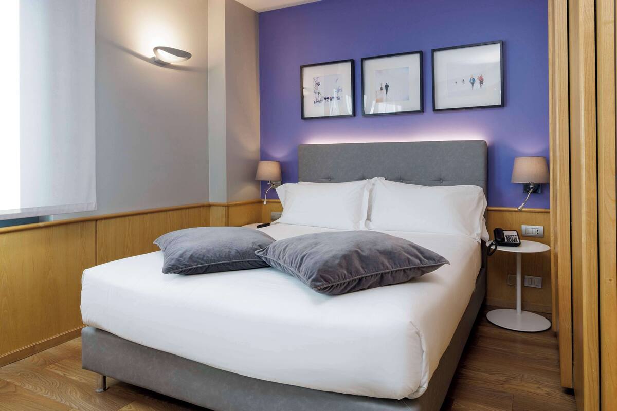 A modern hotel room with a large bed, two bedside tables, and a wall-mounted lamp.
