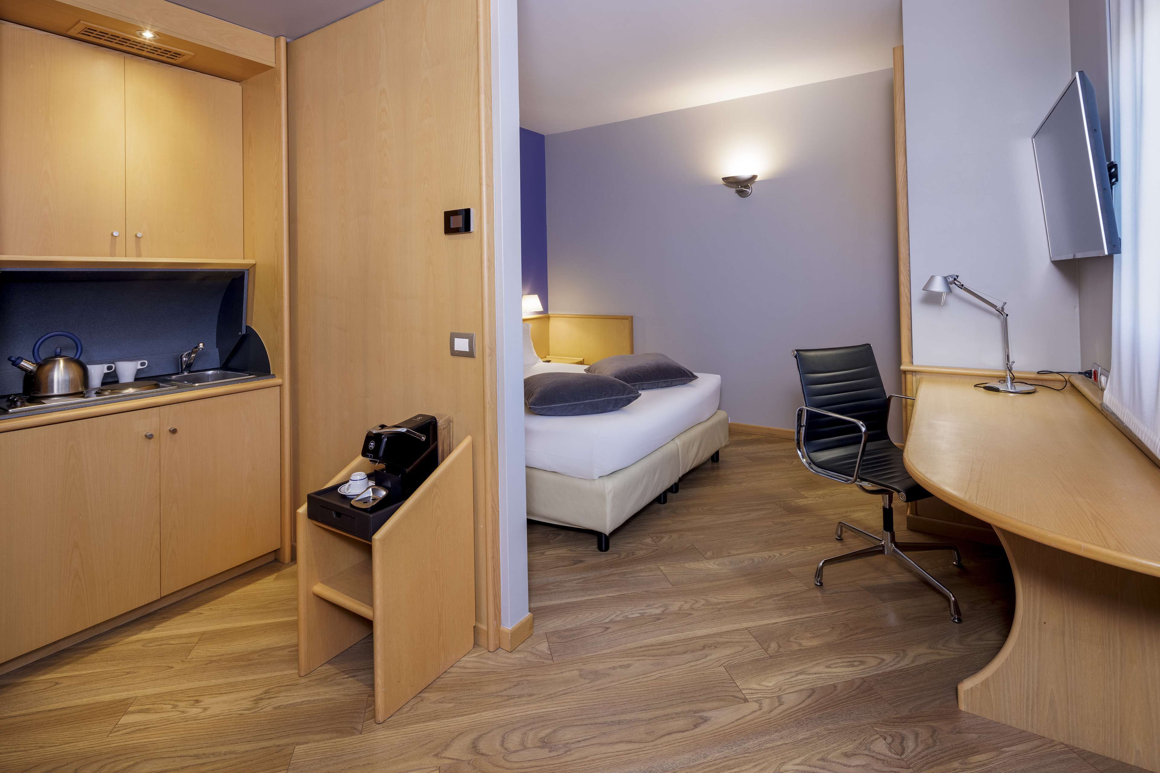 A compact hotel room with a kitchenette, a bed with pillows, a desk with a chair, and a TV mounted on the wall.