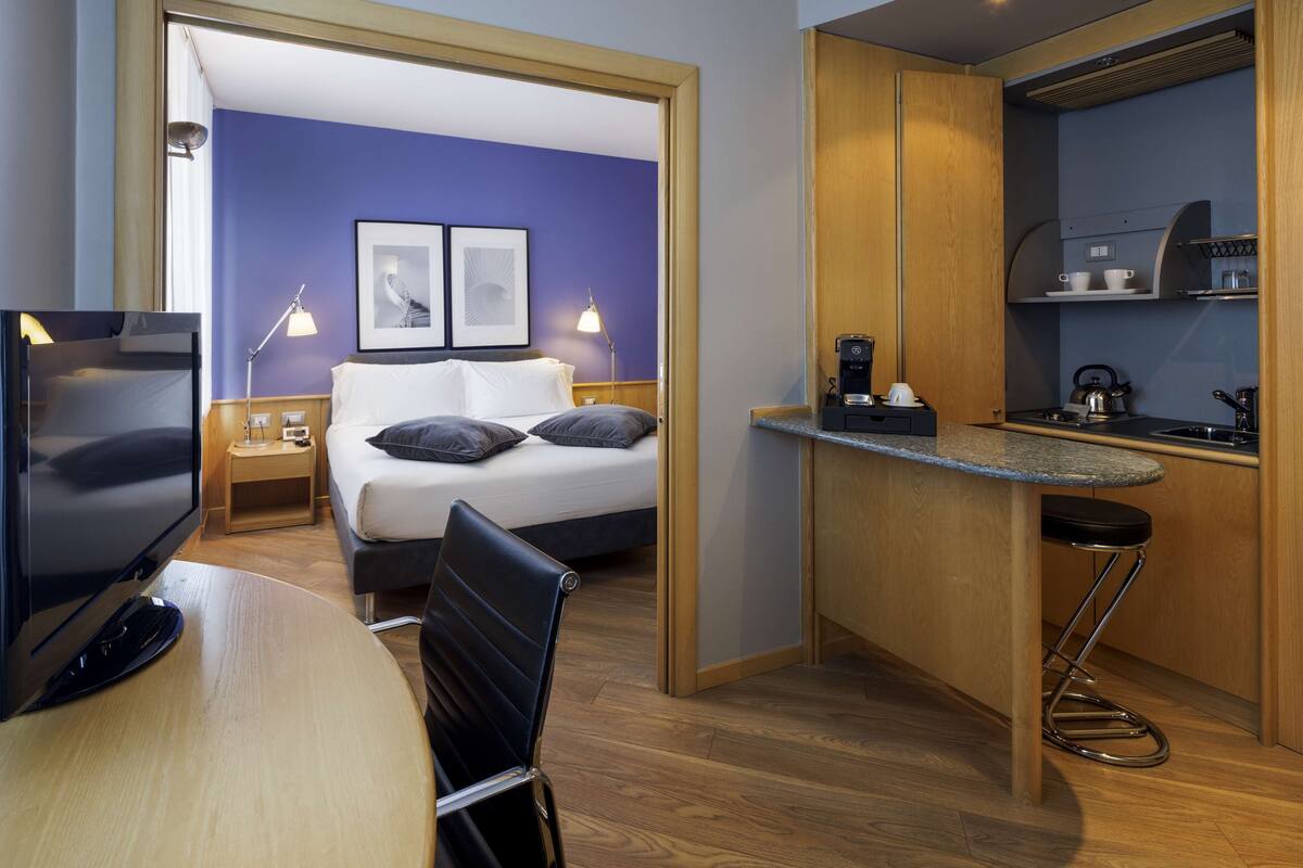A compact hotel room with a bed, a desk, a chair, a TV, a coffee maker, and a small kitchenette.