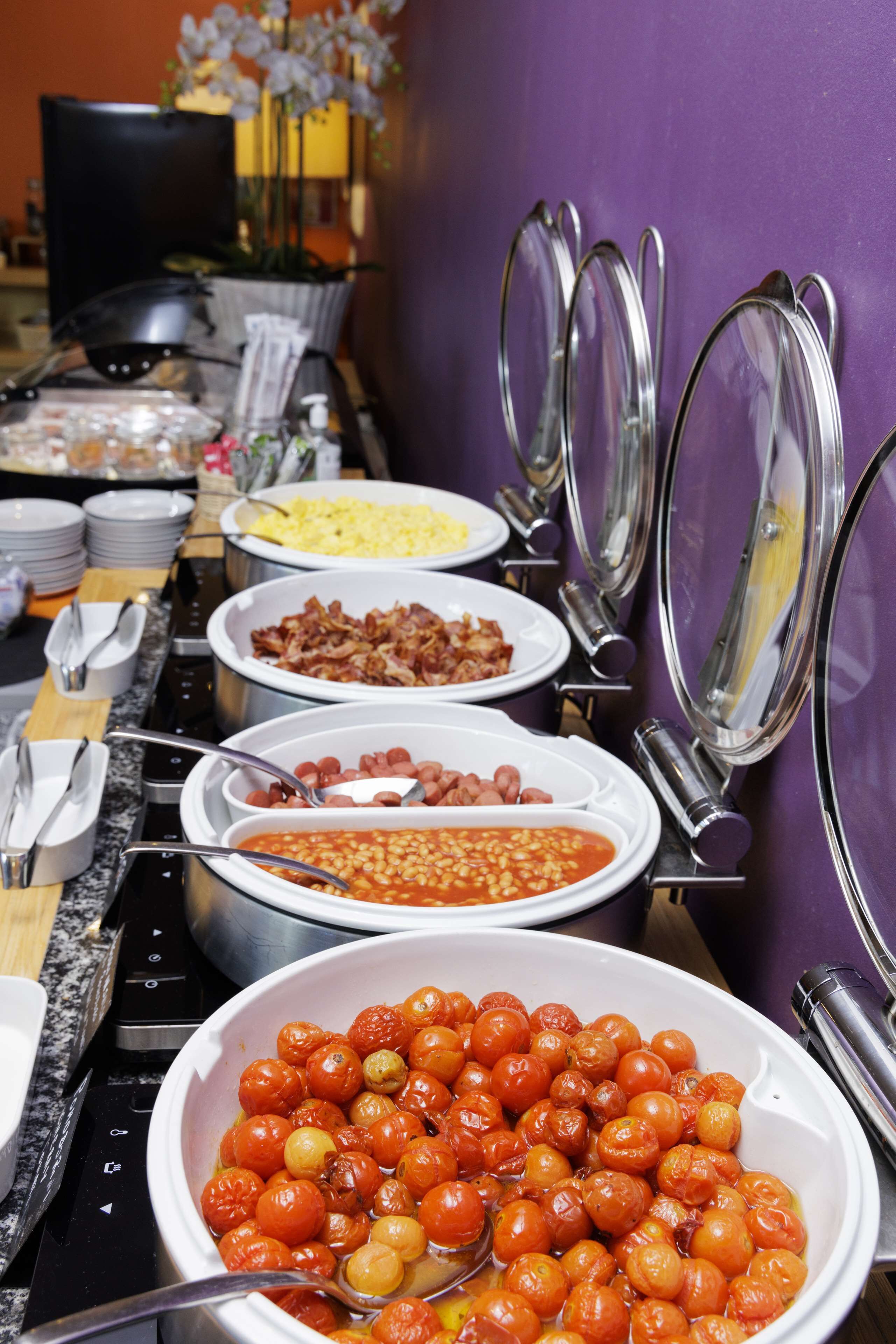 A buffet with various dishes including beans, bacon, and a large bowl of cherry tomatoes.