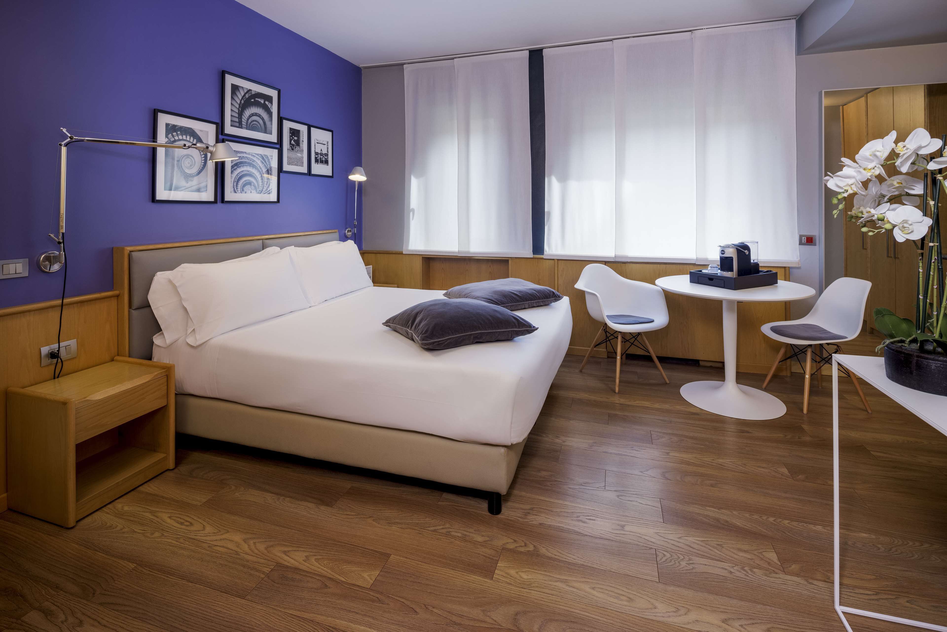 A modern hotel room with a bed, bedside table, lamp, and a small desk with chairs.