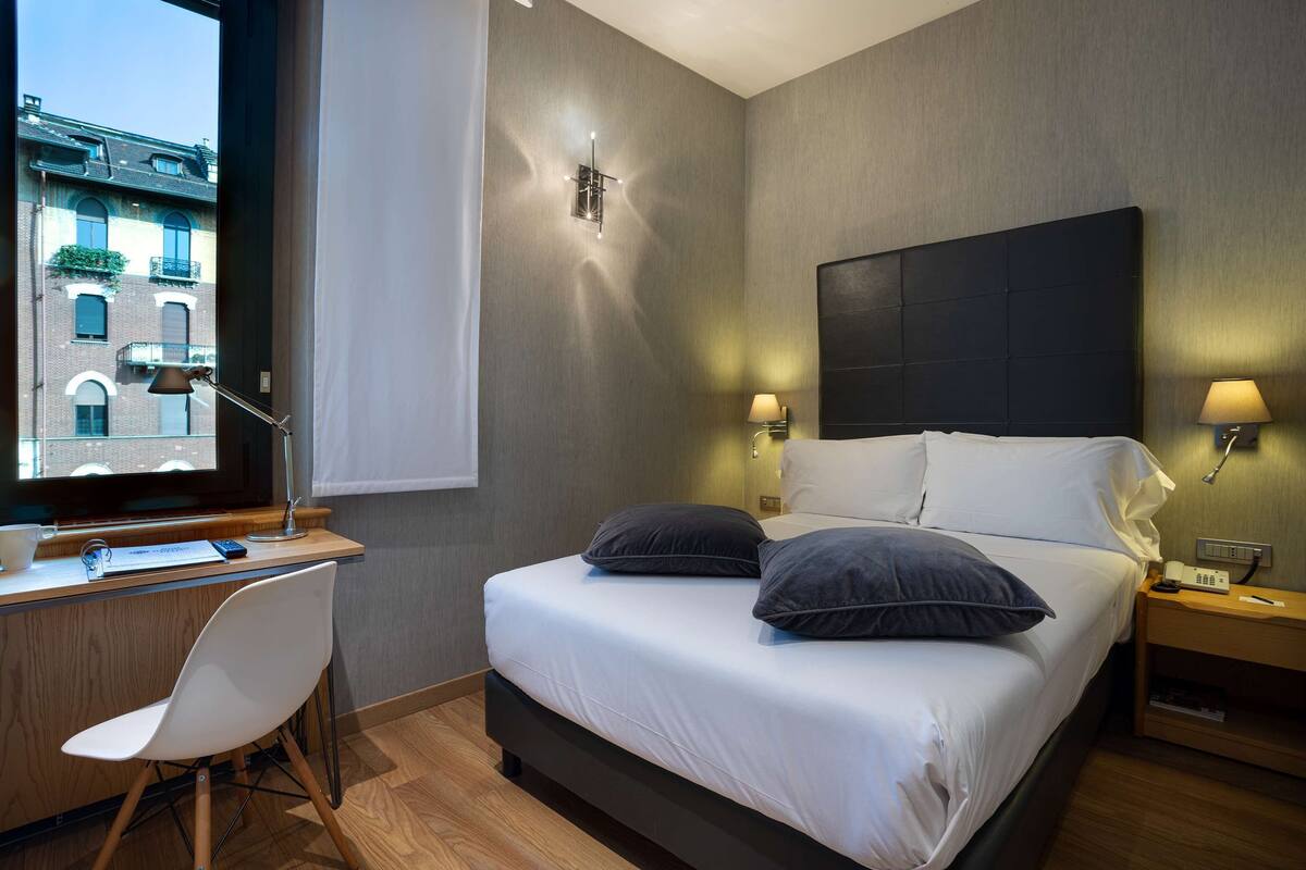 A modern hotel room with a large bed, bedside lamps, a desk with a chair, and a view of buildings outside the window.
