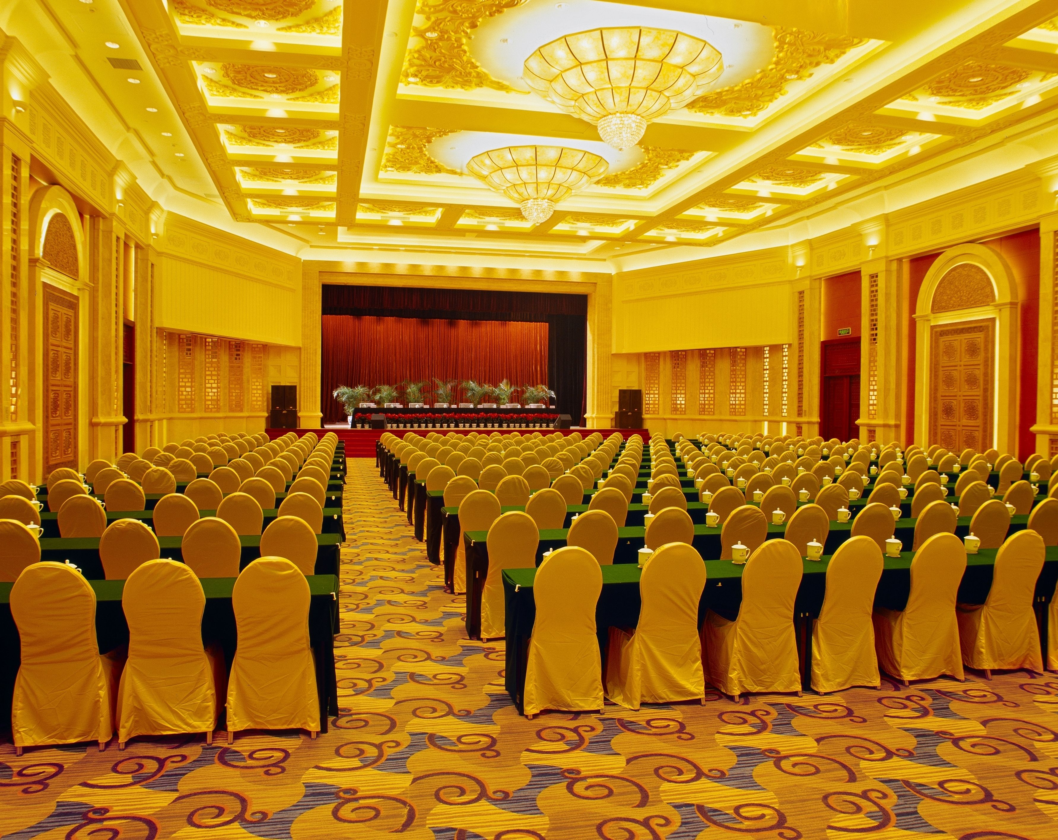 A conference room with rows of tables and chairs, a stage with a red curtain, and ornate ceiling details.