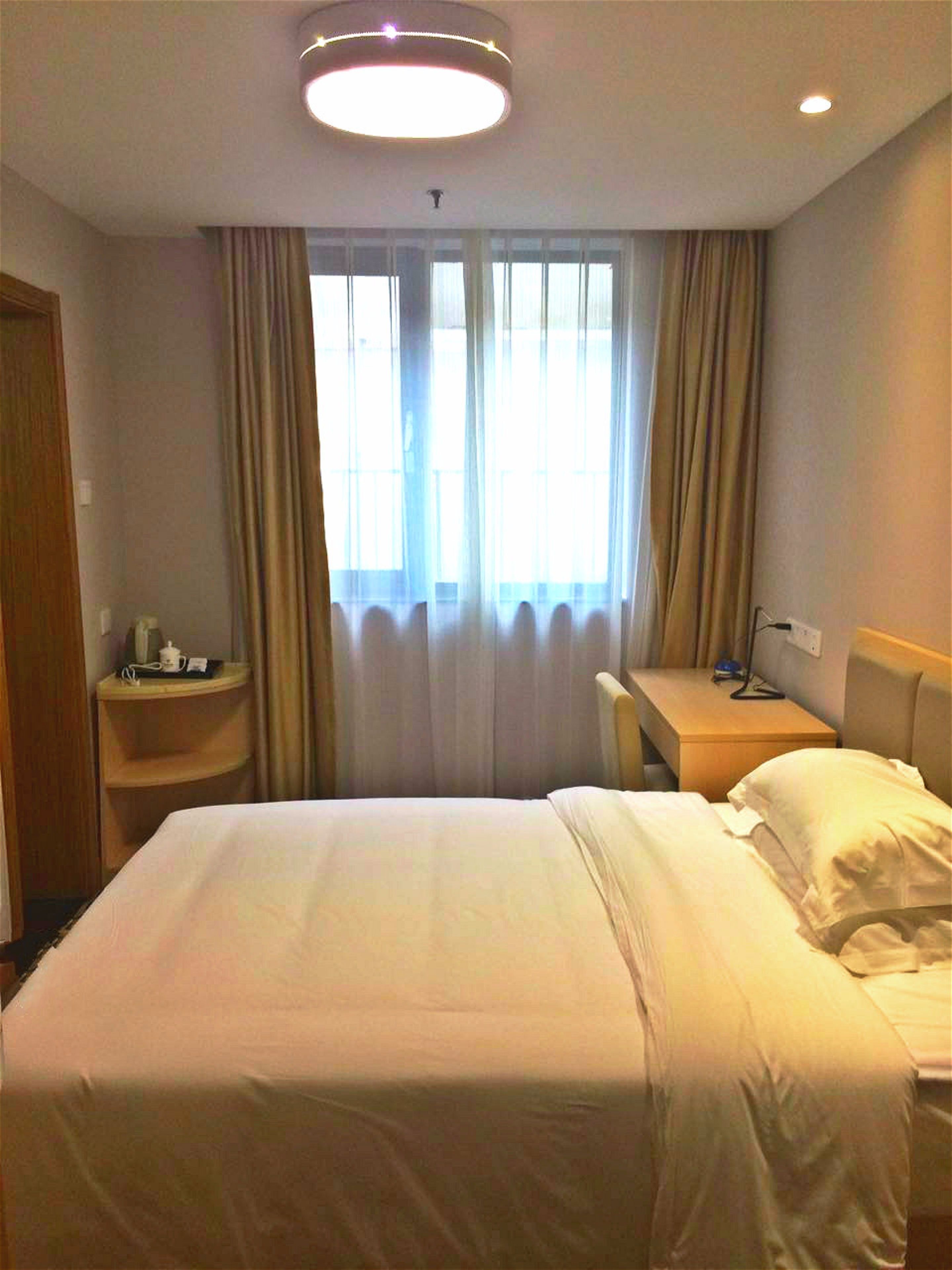 Ett hotellrum med en stor säng, ett skrivbord och en stol. Det finns ett fönster med gardiner och en taklampa.