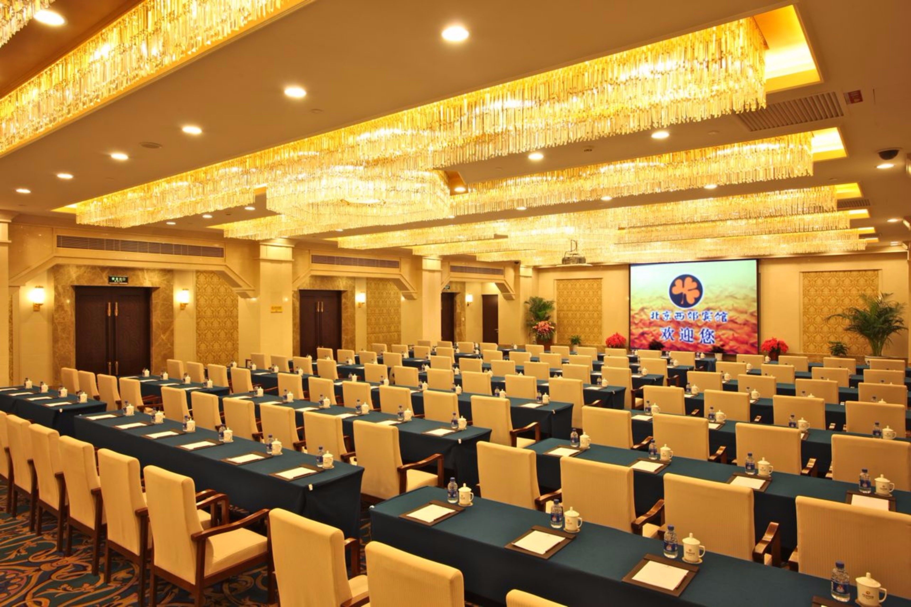 A large conference room with rows of tables and chairs, a large screen, and chandeliers.