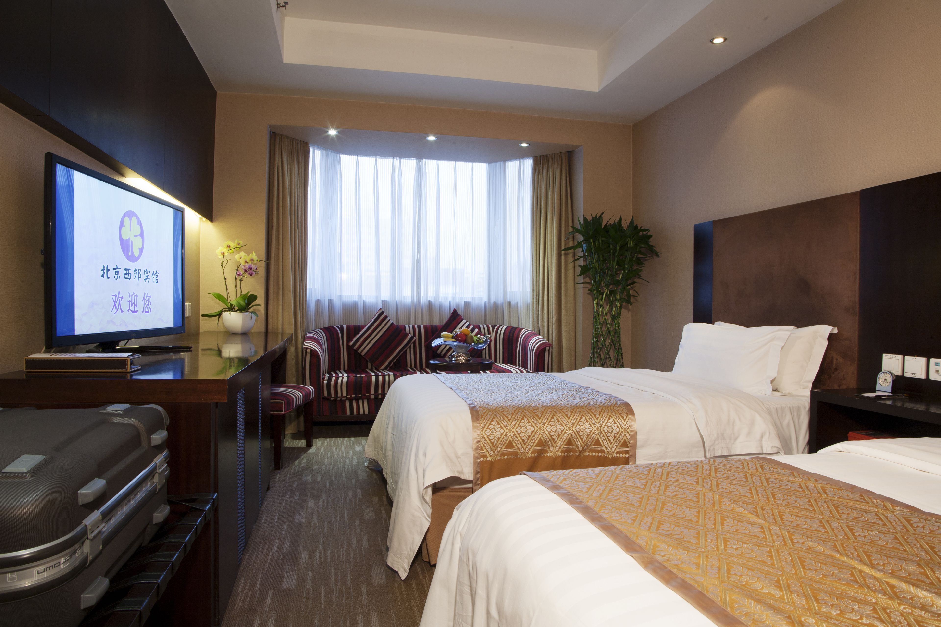 A hotel room with two beds, a desk with a television, a seating area with a sofa and chairs, and a large window with curtains.