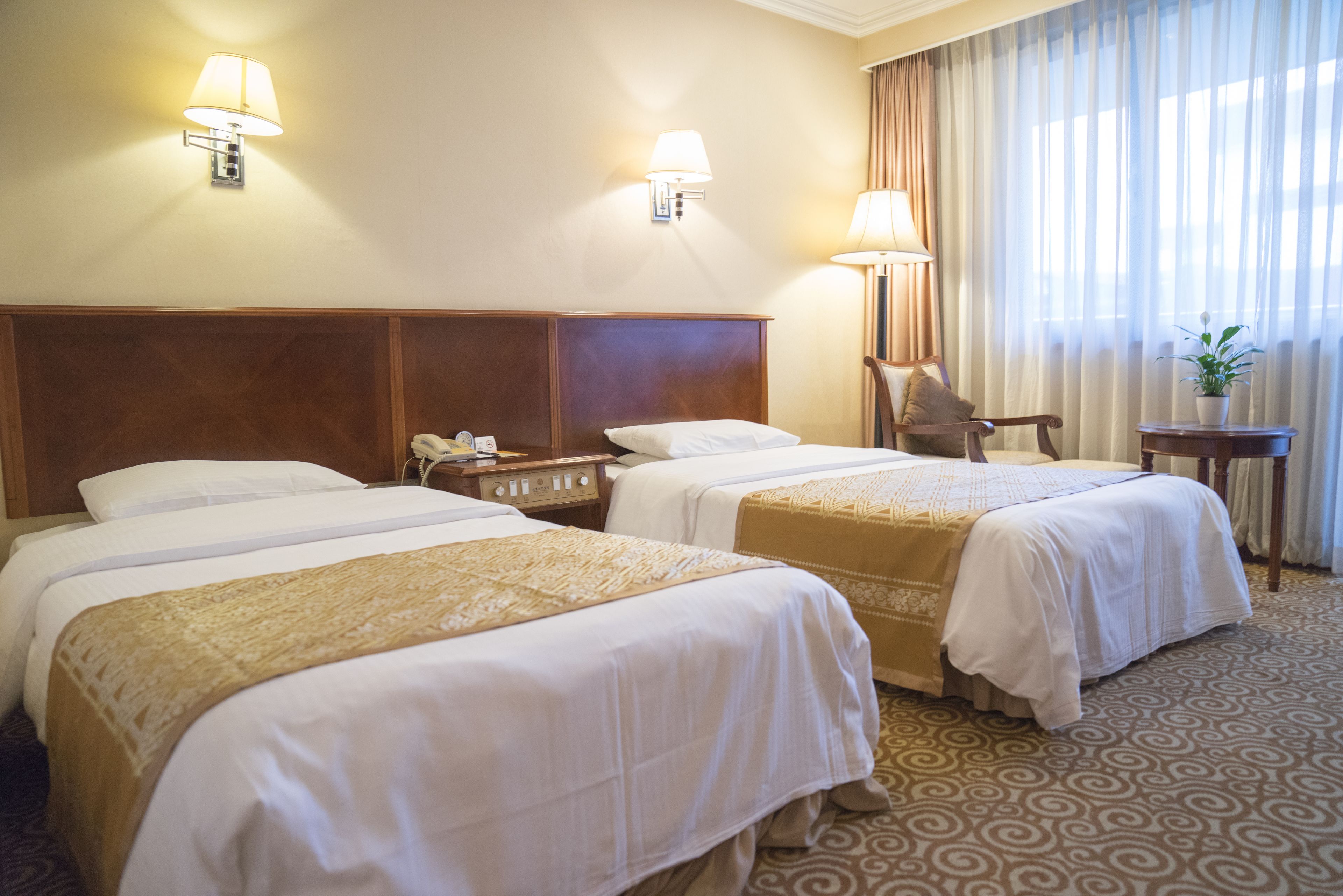 A hotel room with two beds, a wooden headboard, a chair, a small table, and a lamp.