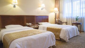 A hotel room with two beds, a wooden headboard, a chair, a small table, and a lamp.