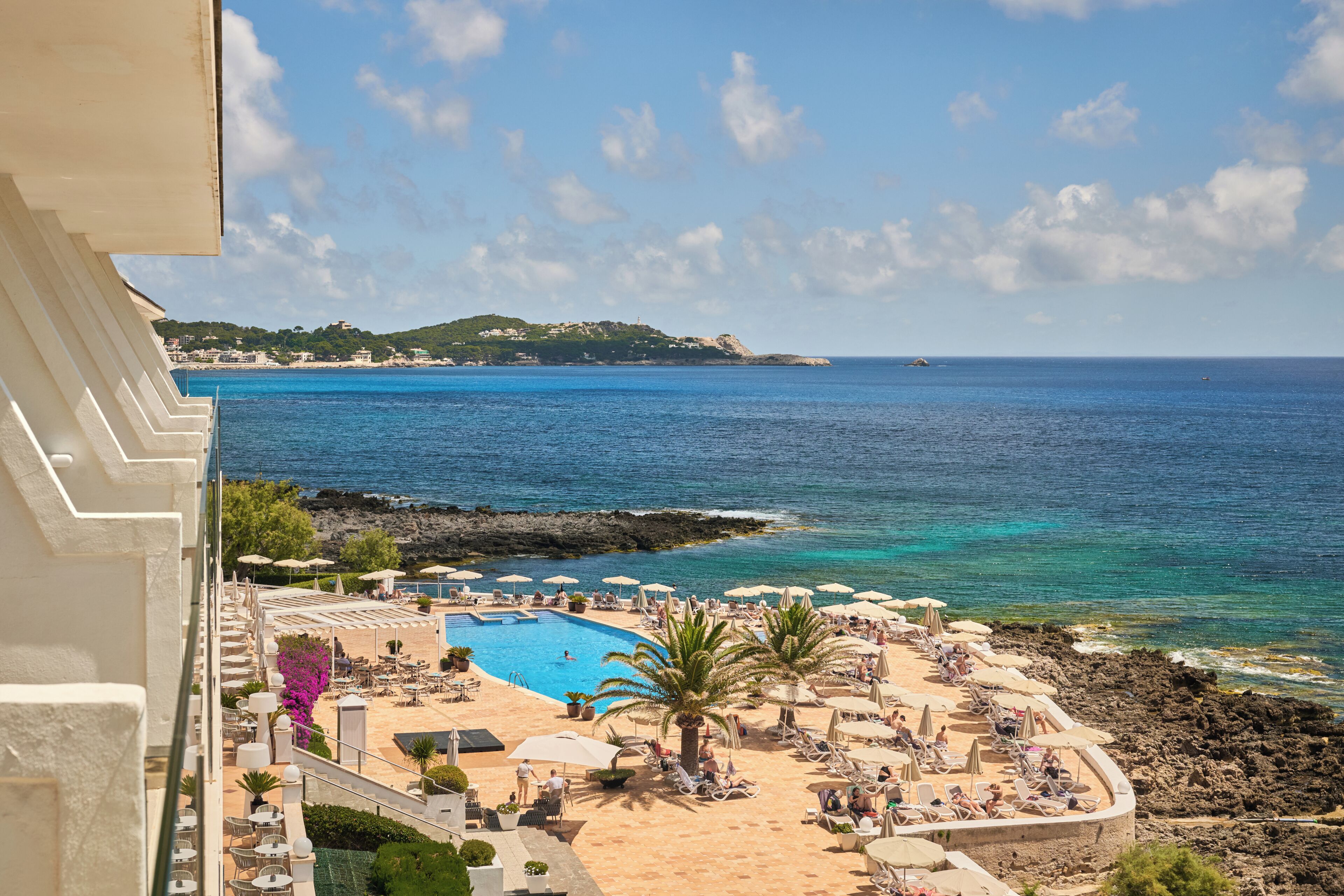 A resort with a pool, beach chairs, umbrellas, and a view of the sea and distant hills.