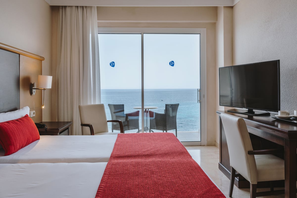A hotel room with two beds, a TV, a desk, and a view of the sea.