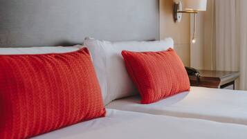 Habitación de hotel con una cama, dos almohadas rojas, una almohada blanca, una mesita de noche con una lámpara y un aplique de pared.