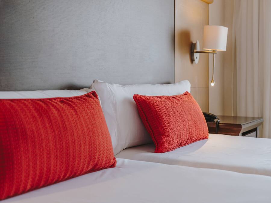 Habitación de hotel con una cama, dos almohadas rojas, una almohada blanca, una mesita de noche con una lámpara y un aplique de pared.