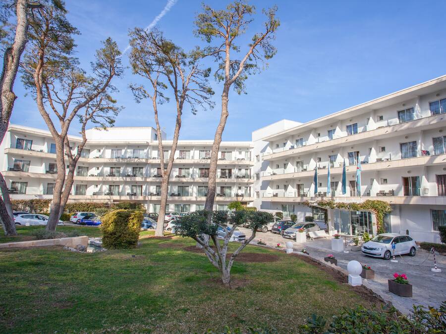 A multi-story hotel with balconies, a parking area, and a well-maintained lawn with trees.