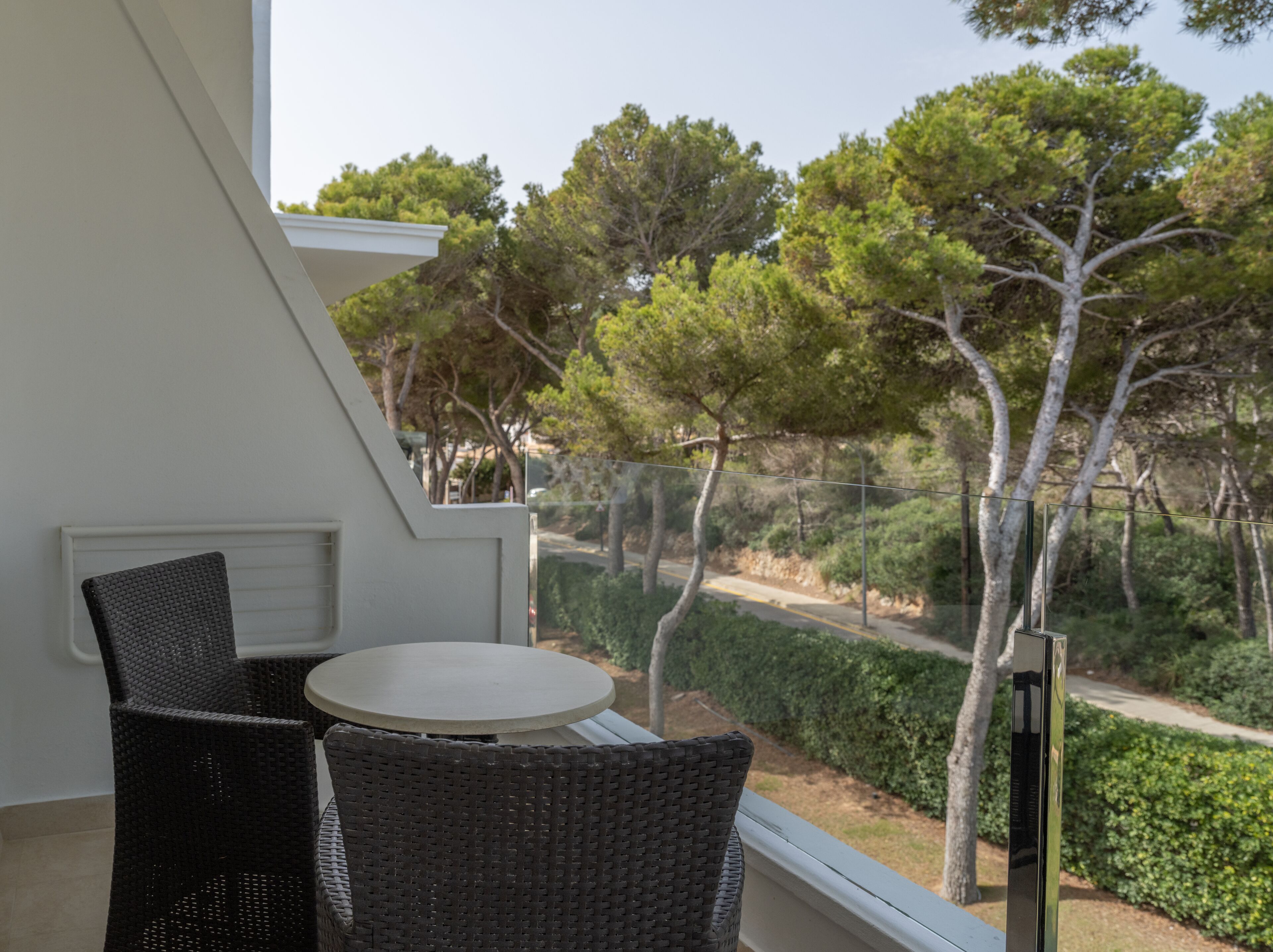 A balcony with wicker chairs and a small table, overlooking a landscaped area with trees and a pathway.