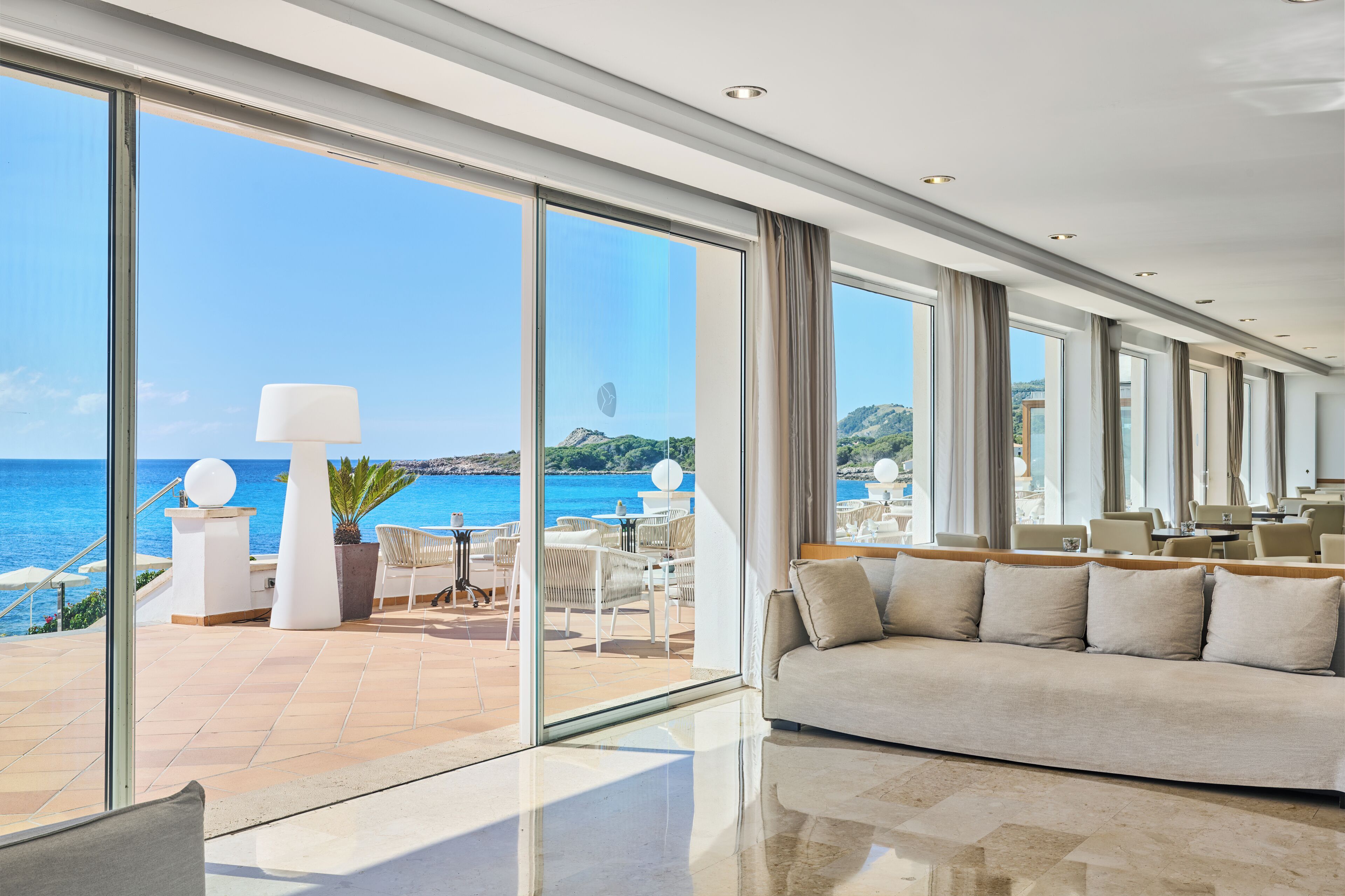 A modern hotel room with large windows, a balcony with outdoor seating, and a view of the sea.