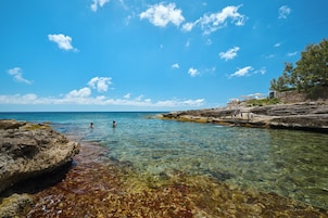 Escena costera con agua cristalina, costa rocosa y algunas personas nadando.