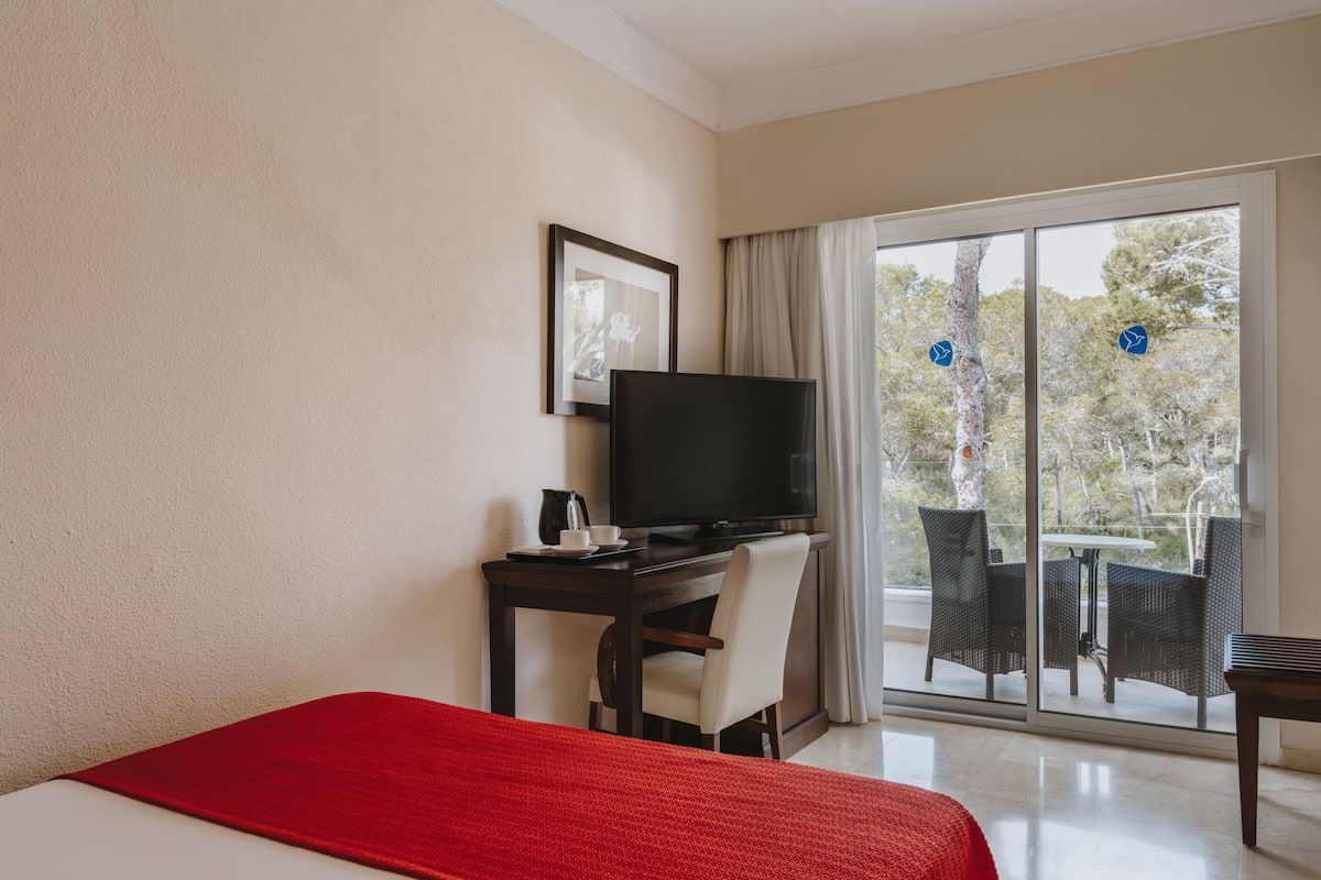 A hotel room with a bed, a desk with a TV, a chair, and a balcony with outdoor furniture.