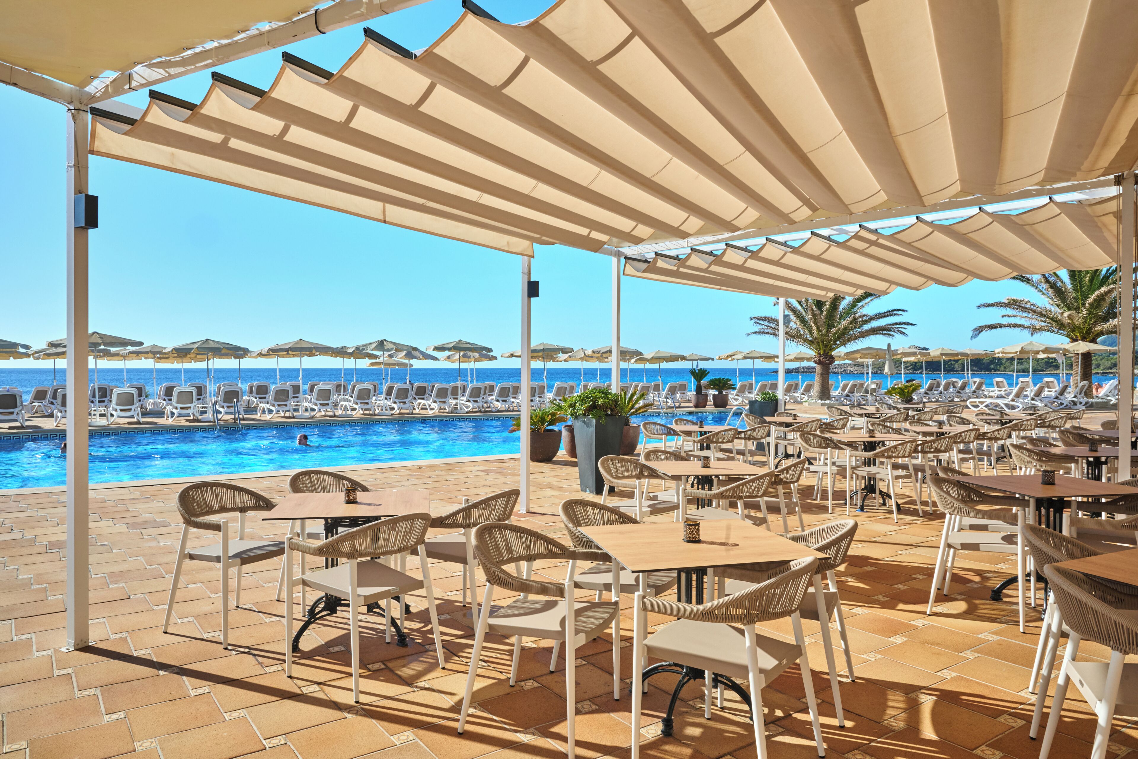 A poolside area with lounge chairs and tables, a clear view of the sea, and a covered seating area with umbrellas.