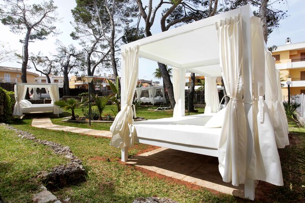 Una cama blanca al aire libre con cortinas, rodeada de un jardín y árboles.