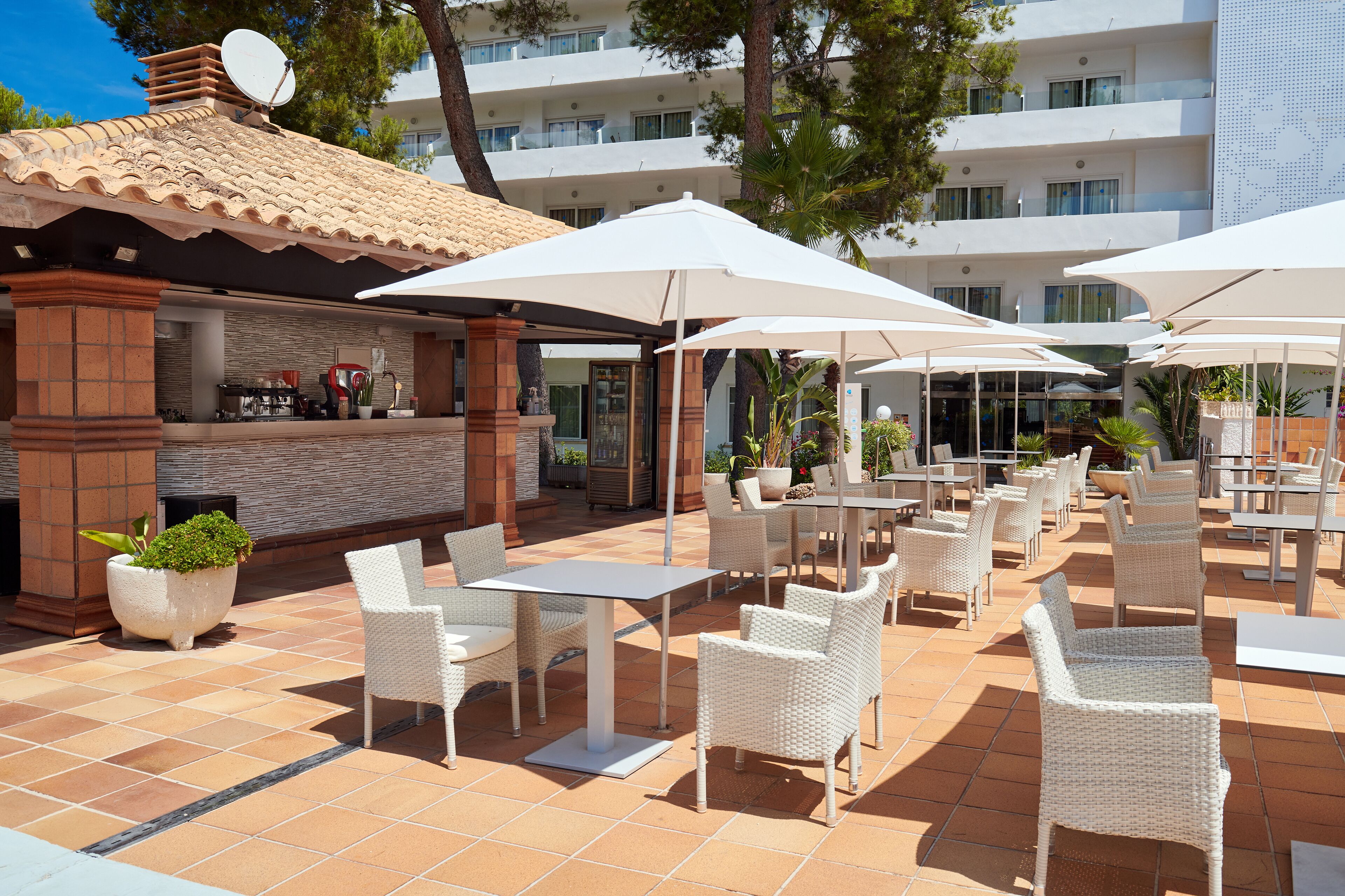 An outdoor patio with white wicker furniture, a bar area, and a poolside view of a hotel building.