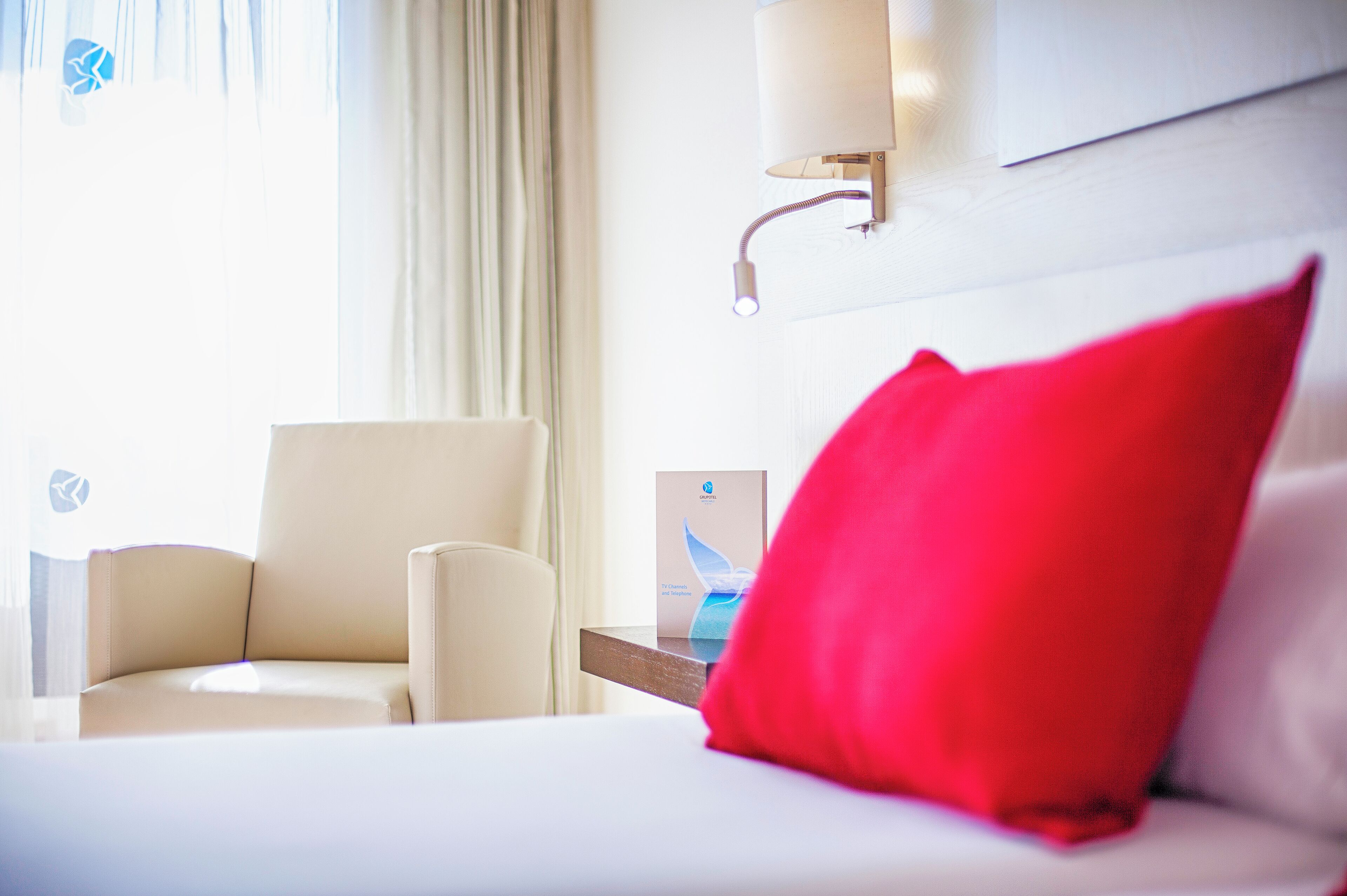 Ein Hotelzimmer mit Bett, Stuhl, Nachttisch, Lampe und einem Fenster mit Vorhängen.