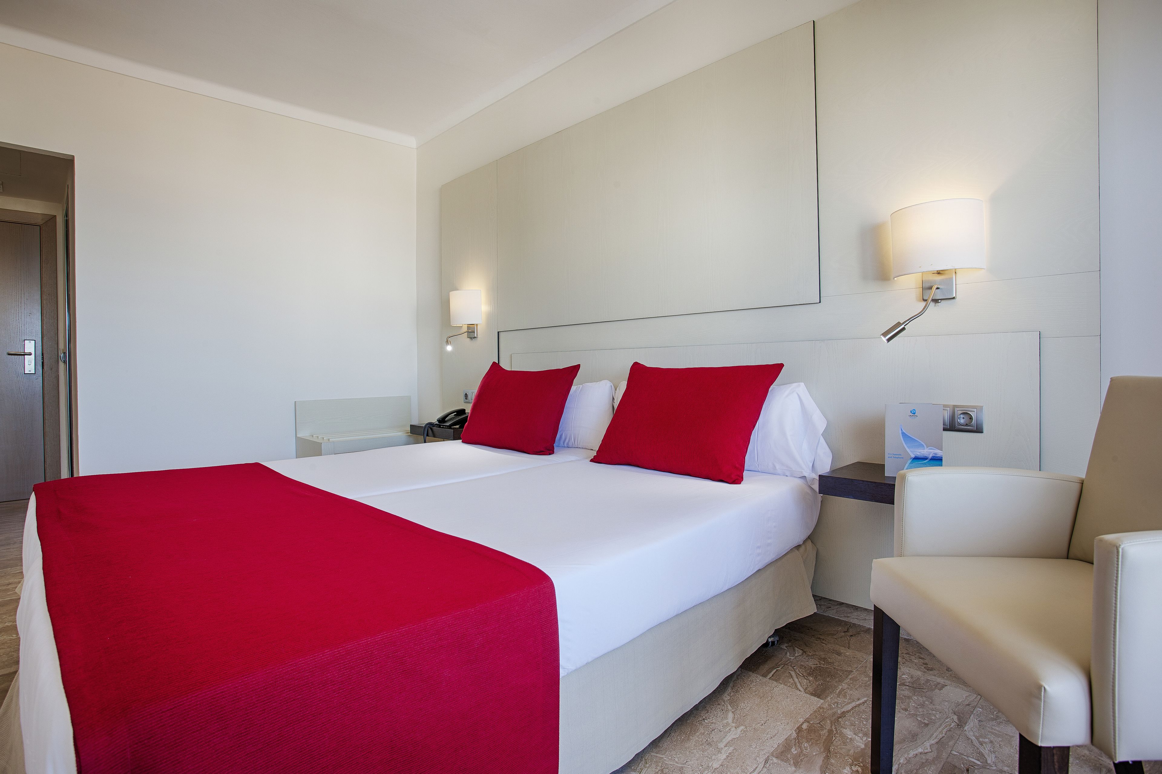 Quarto de hotel com uma cama grande, duas almofadas vermelhas, um edredom branco, uma mesa de cabeceira com abajur e uma cadeira.