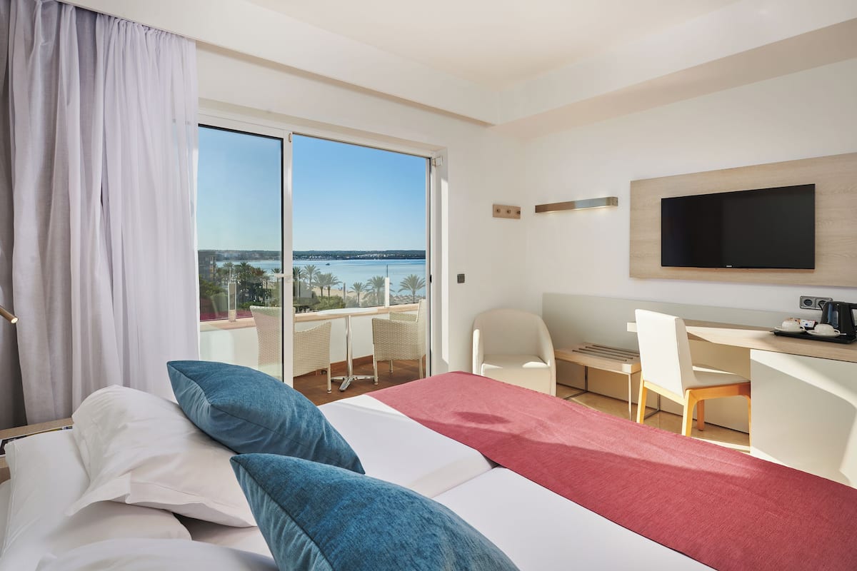 A hotel room with a bed, a desk, a chair, a TV, and a view of the sea.