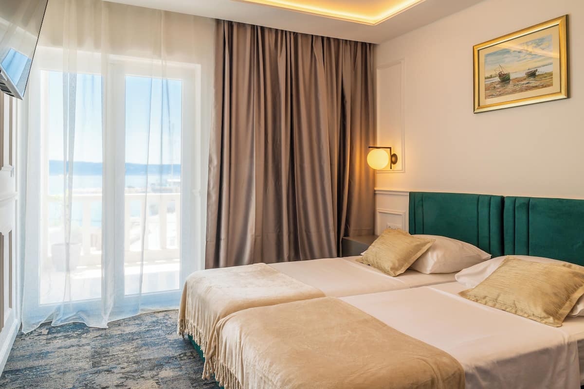 Quarto de hotel com duas camas, uma janela grande com cortinas leves e vista para o mar, e um quadro na parede.