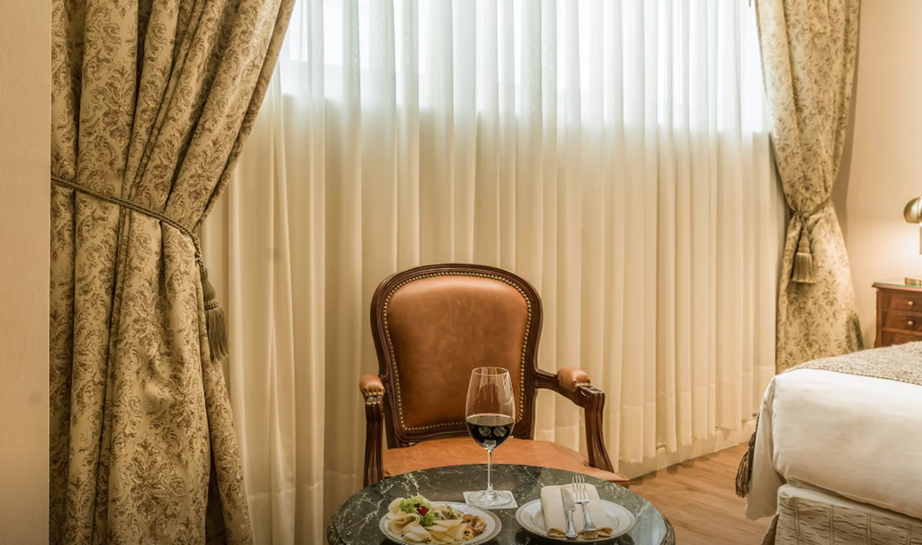 Una habitación con una mesa de comedor preparada para dos personas, una silla, una copa de vino tinto y una cama con una colcha blanca.