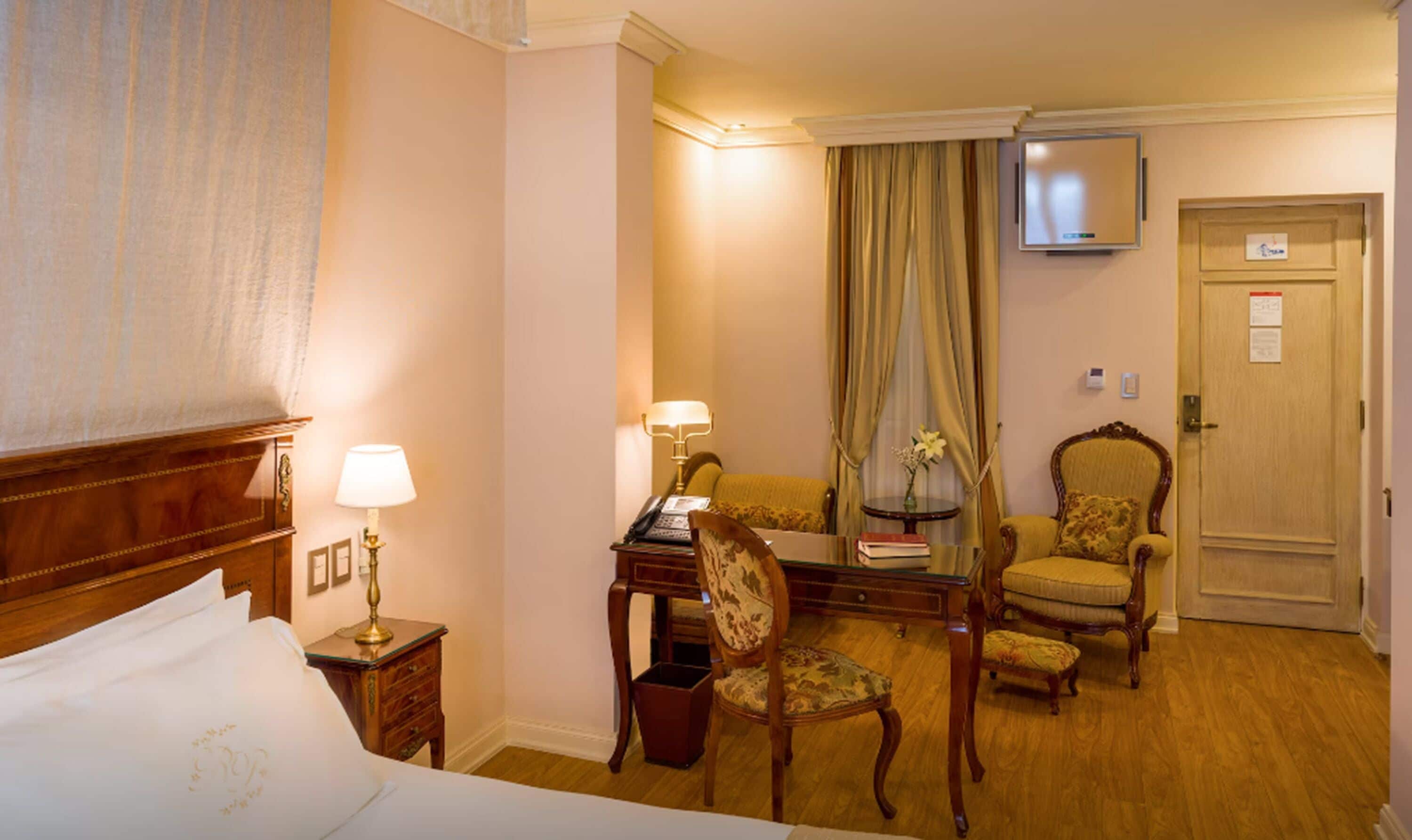 Een hotelkamer met een bed, een bureau met een lamp, twee stoelen, een televisie en een deur.