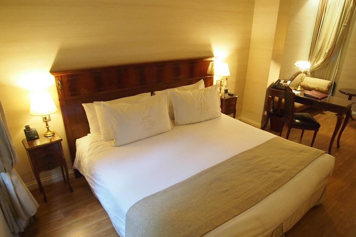 A hotel room with a large bed, two bedside tables with lamps, a desk with a chair, and a window with curtains.