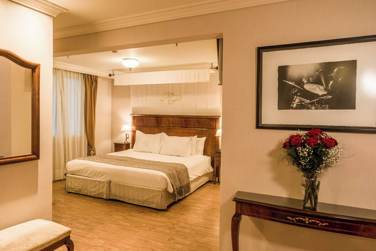 A hotel room with a large bed, a bedside table, a chair, a framed picture, and a vase with flowers.