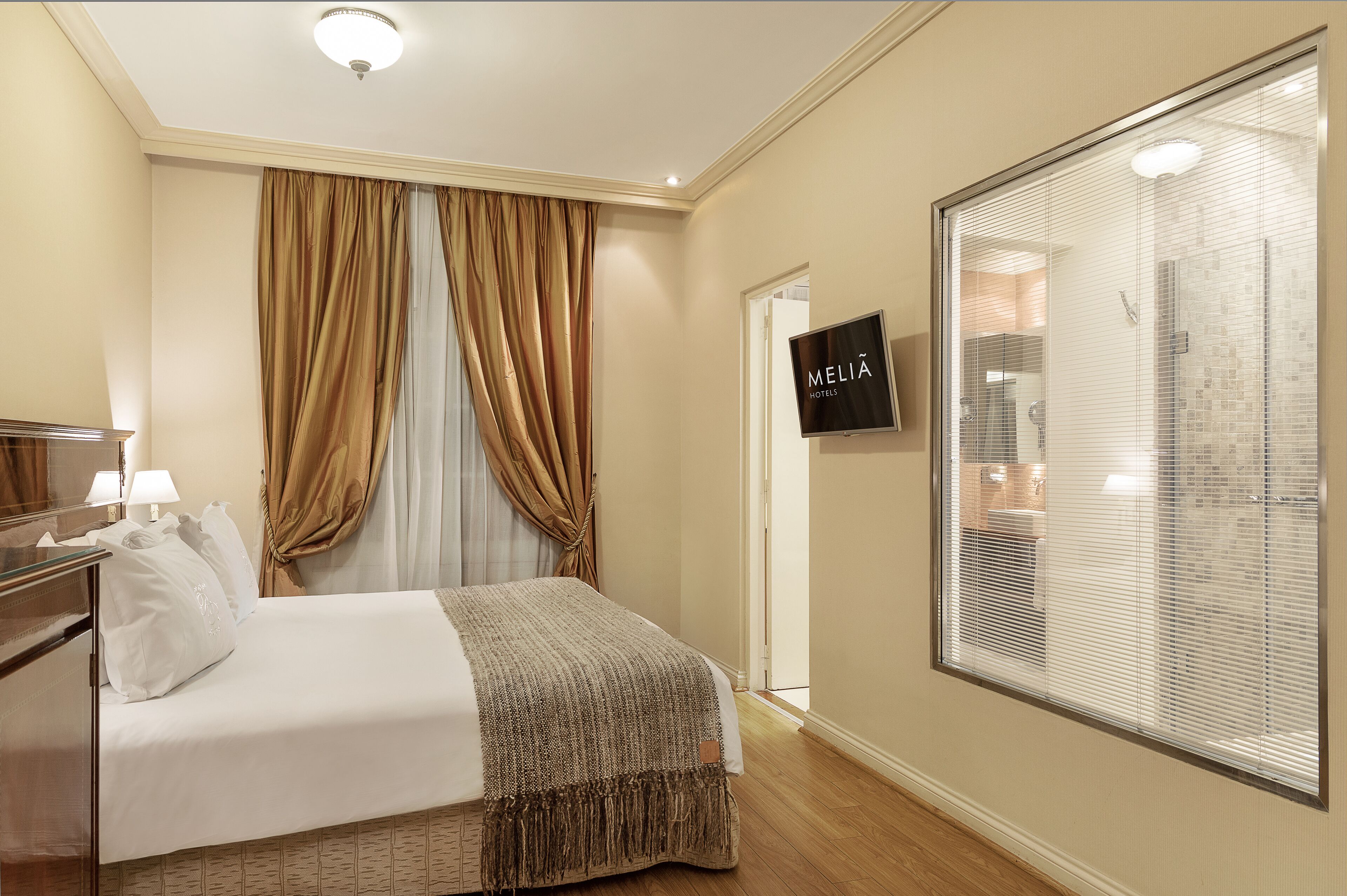Habitación de hotel con cama, televisor y un ventanal grande con persianas.