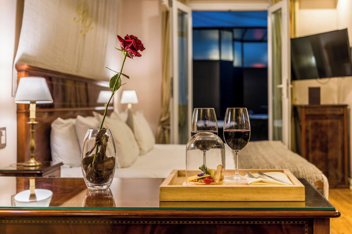 A hotel room with a bed, a bedside table with a vase of flowers, two wine glasses, and a tray with food and cutlery.