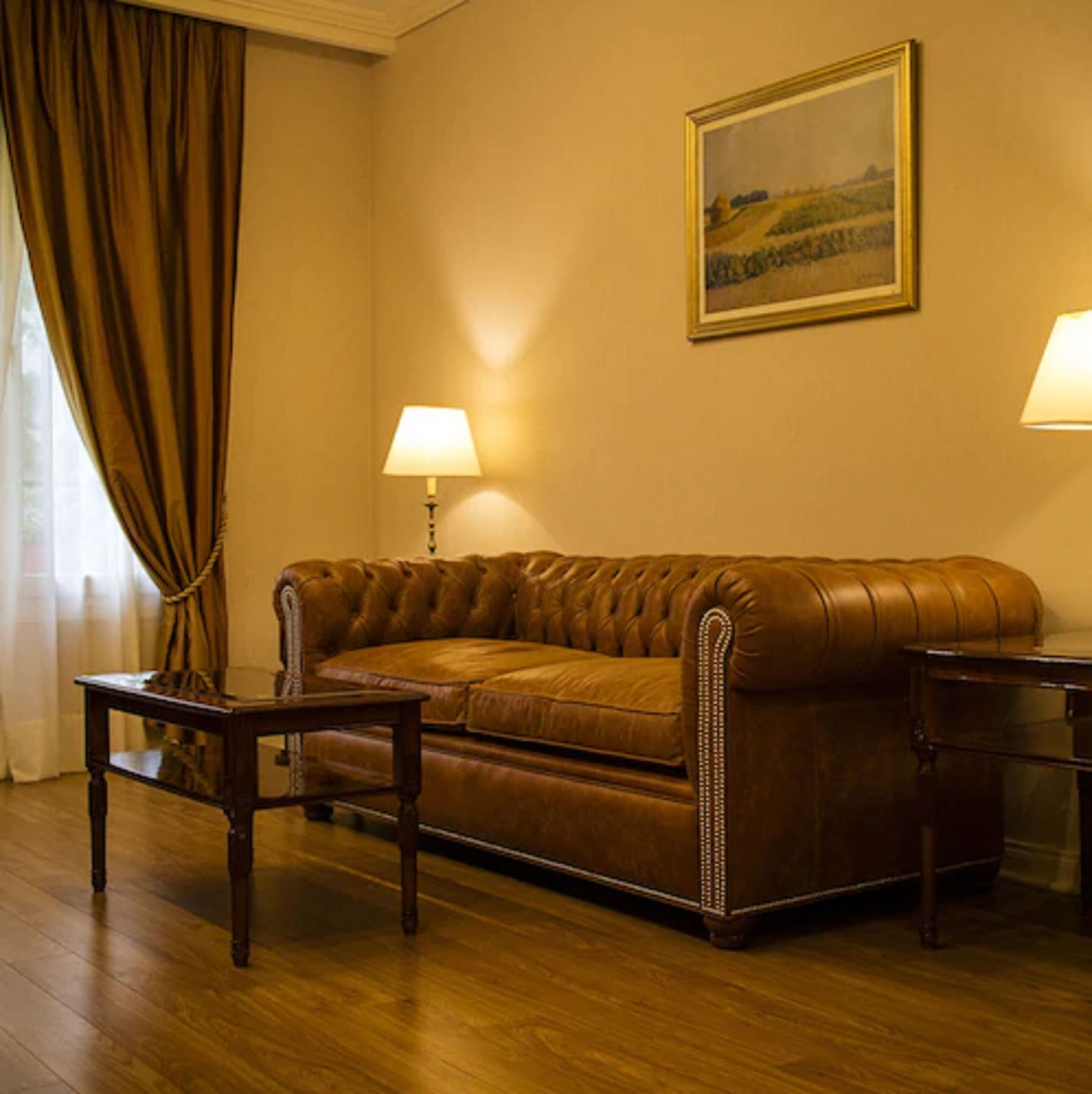 Una habitación con un sofá de cuero marrón, dos mesitas, una lámpara a cada lado, un cuadro en la pared y una ventana con cortinas.