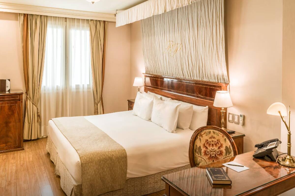 A hotel room with a large bed, two bedside lamps, a wooden desk with a telephone, and a small cabinet.