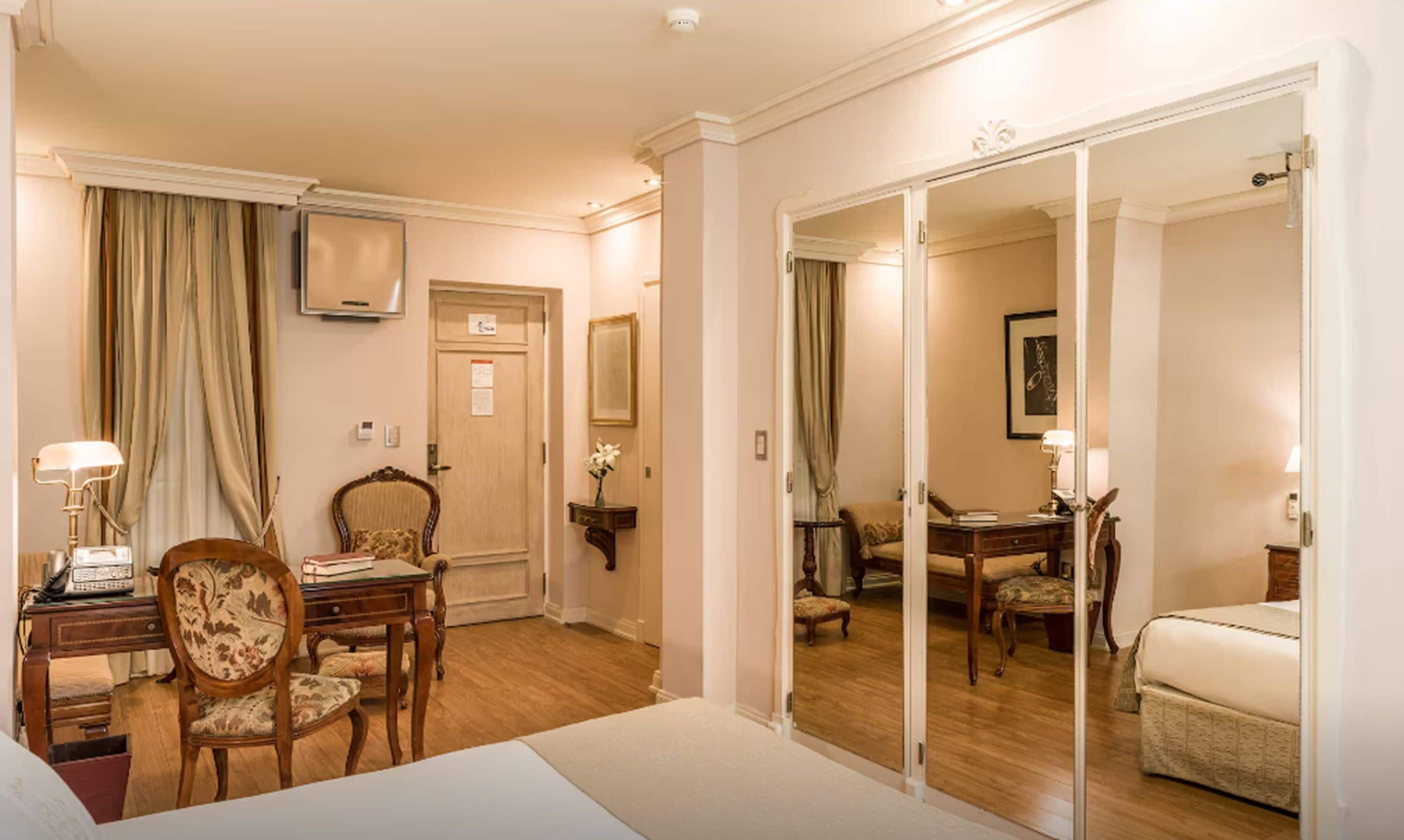 A hotel room with a bed, a desk with a lamp, a chair, a wardrobe, and a mirror.