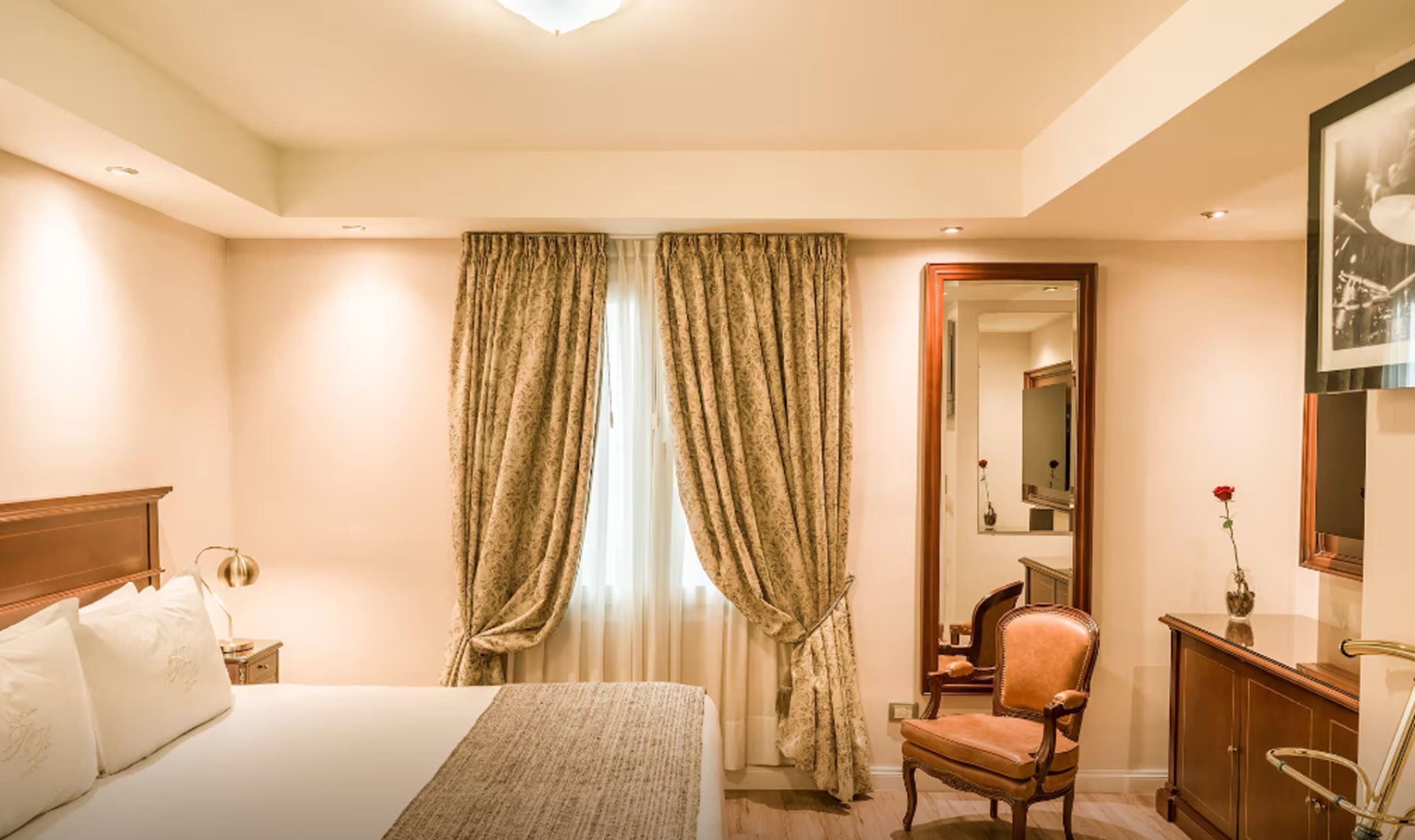 Quarto de hotel com uma cama grande, uma mesa de cabeceira de madeira, uma cadeira, cômoda e um espelho.