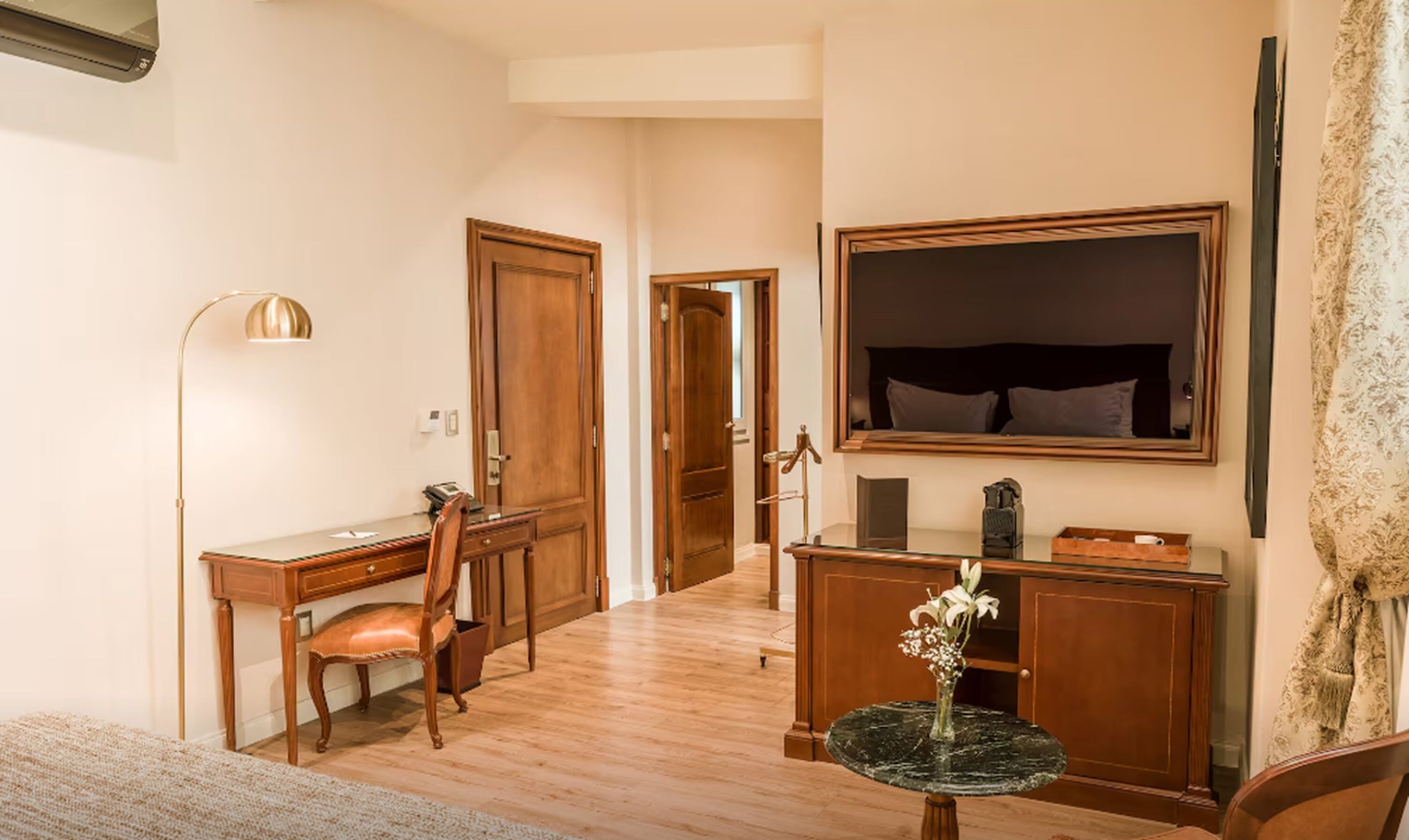A hotel room with a wooden desk, chair, and a round table with a vase. There is a large mirror on the wall and a bed with a headboard.