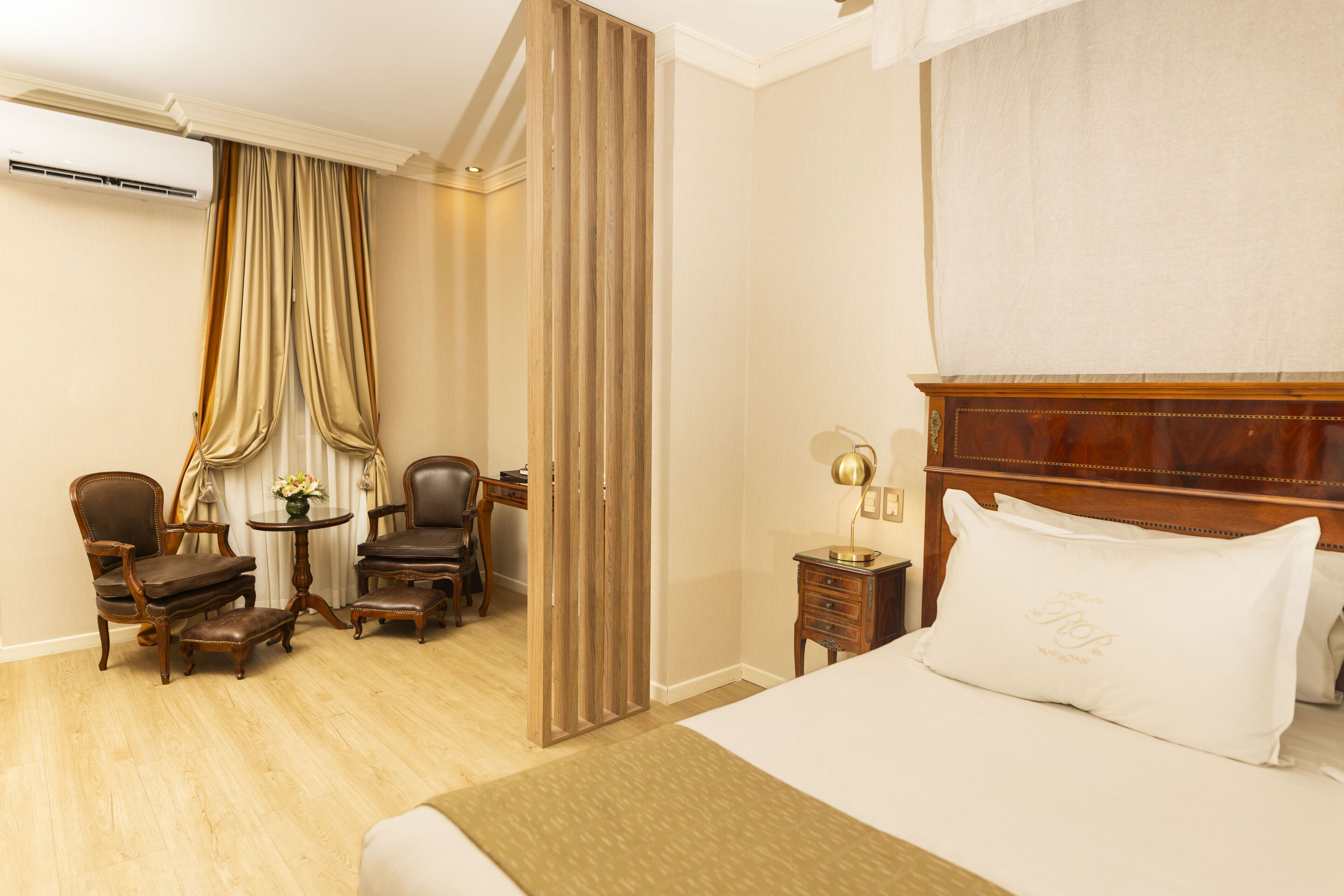 A hotel room with a large bed, a wooden headboard, a desk with two chairs, a small table with a lamp, and a curtain divider.