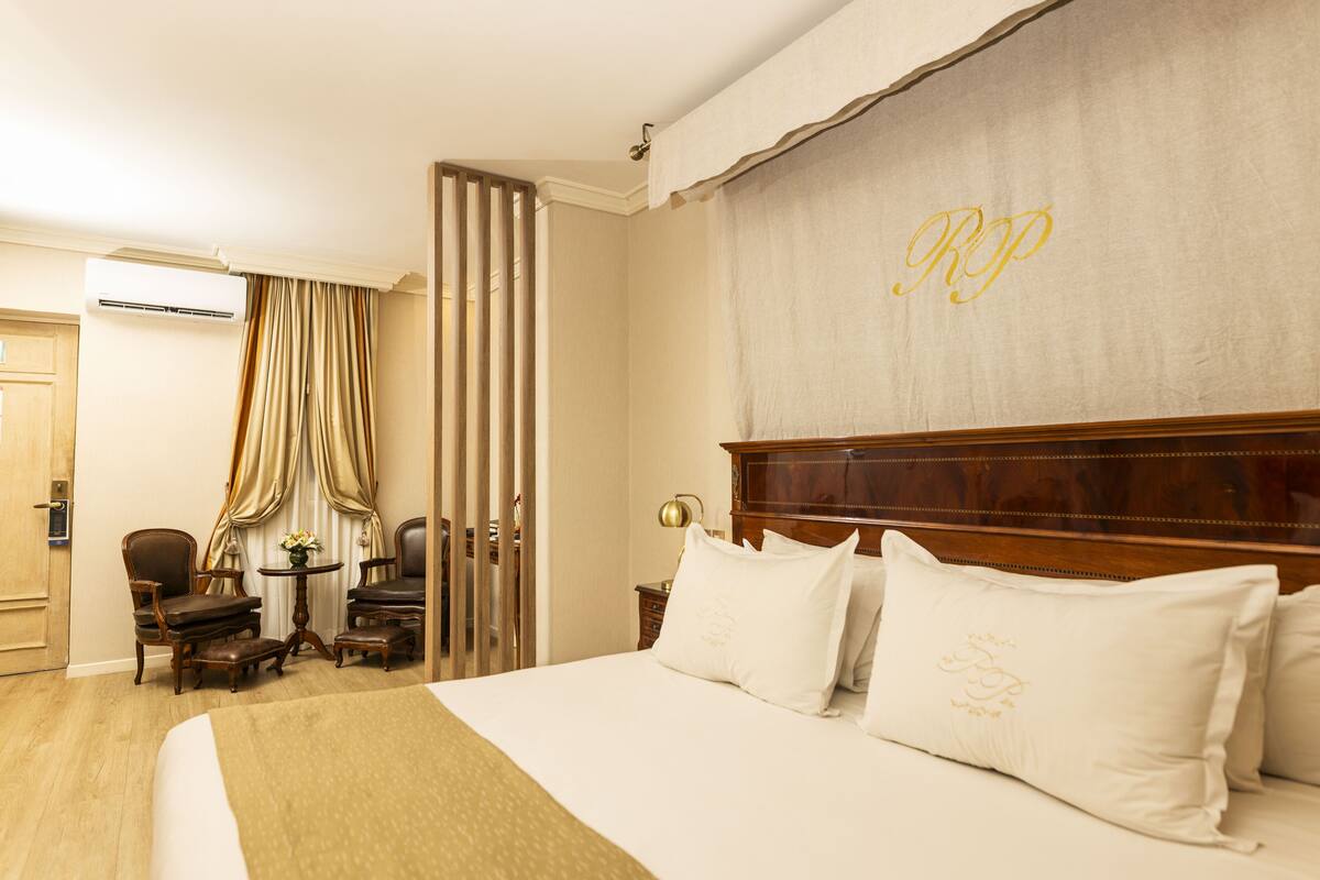 A hotel room with a large bed, a wooden headboard, a desk with a lamp, a chair, and a window with curtains.