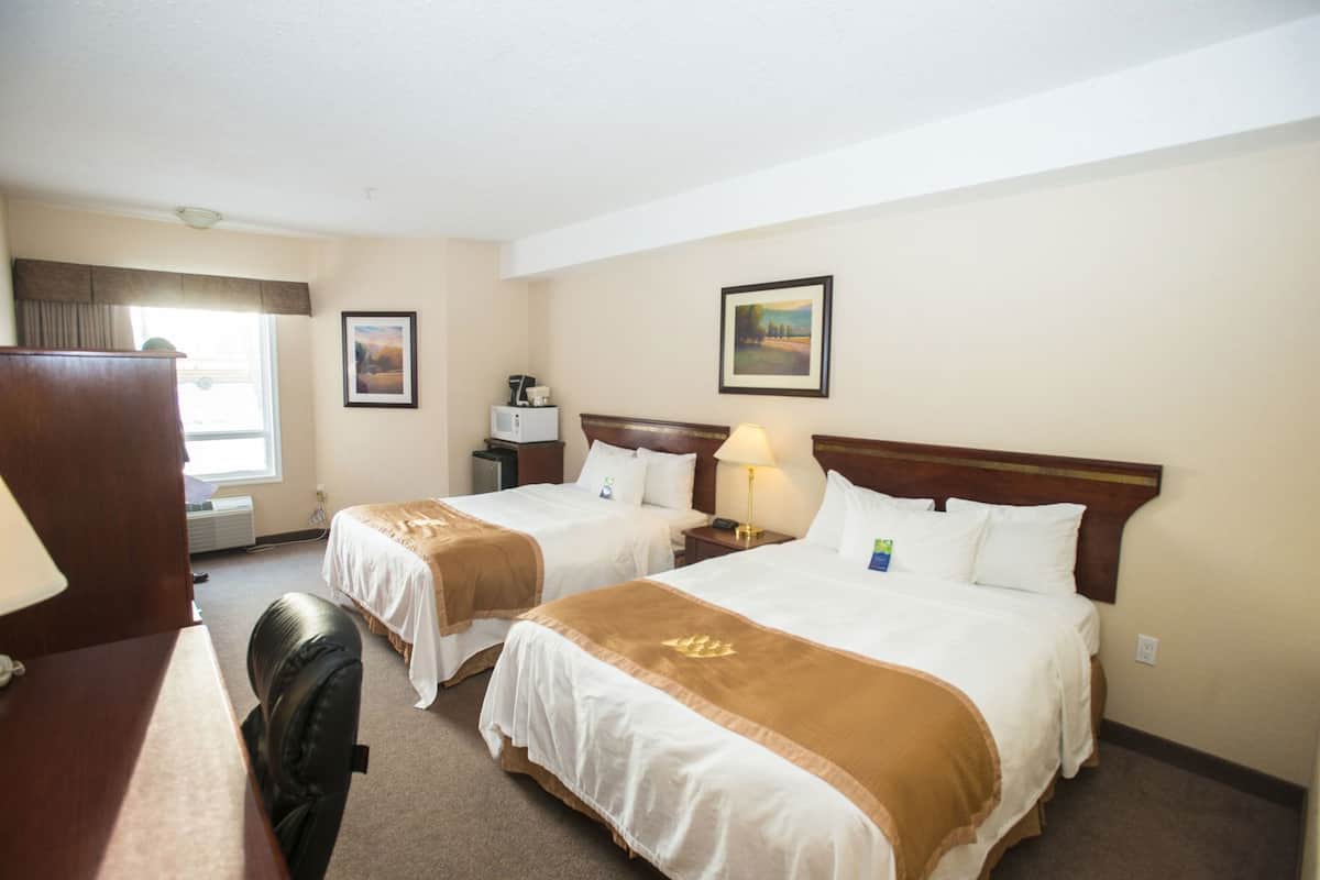A hotel room with two beds, a desk, a chair, a lamp, a microwave, and a painting on the wall.