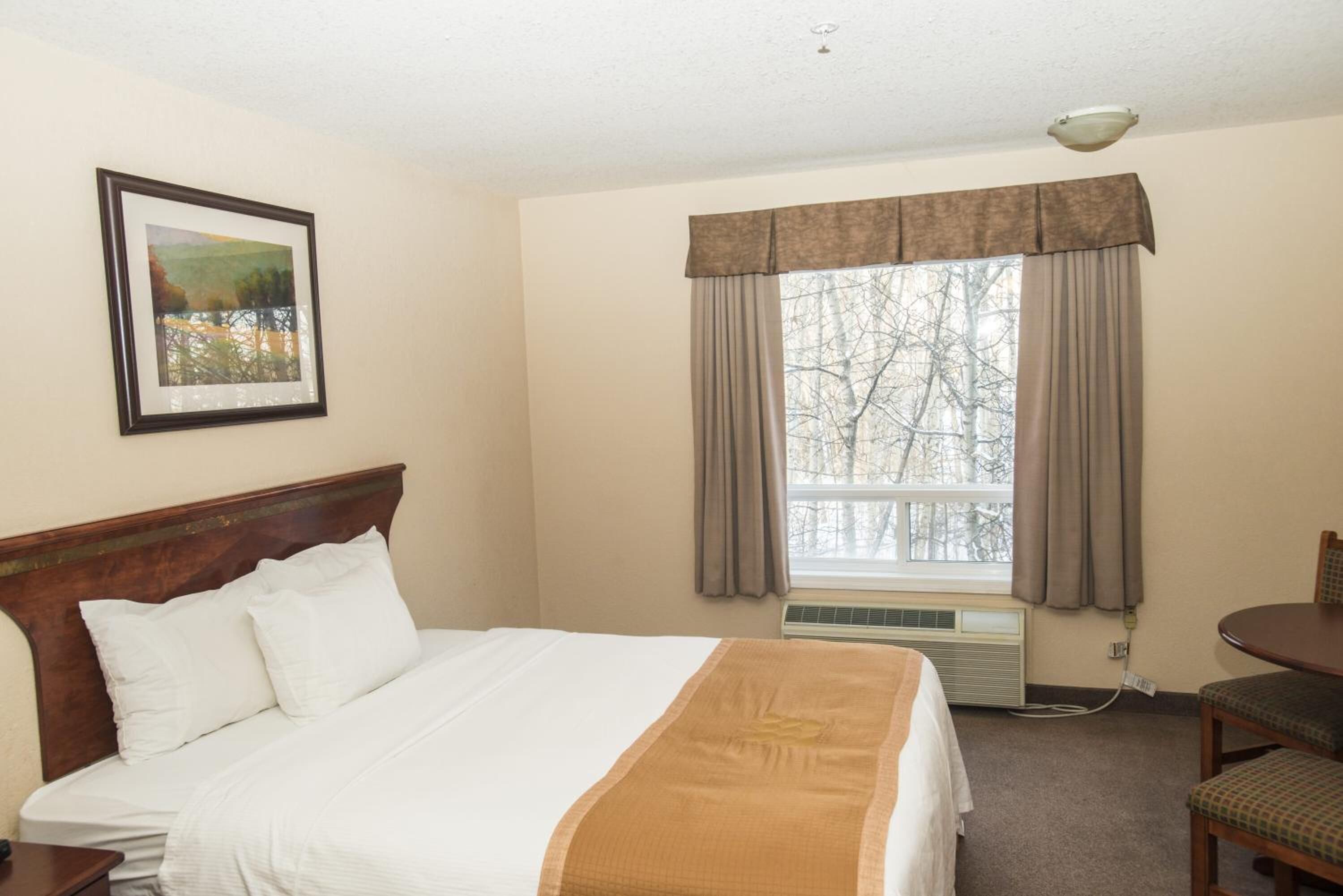 Quarto de hotel com uma cama, um quadro na parede, uma janela com cortinas e uma mesa com cadeiras.