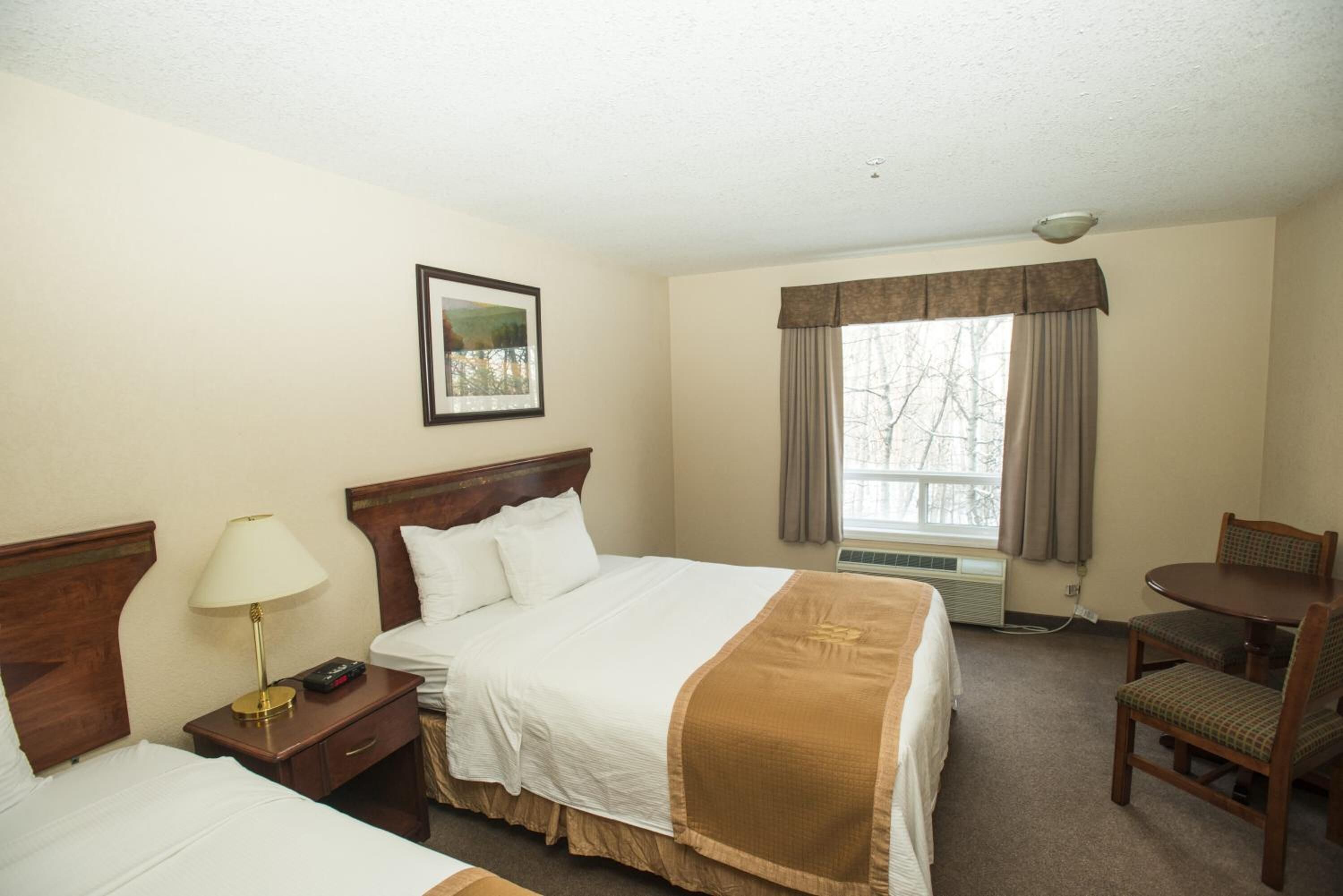 A hotel room with a bed, bedside table with a lamp, a chair, a small table, and a window with curtains.