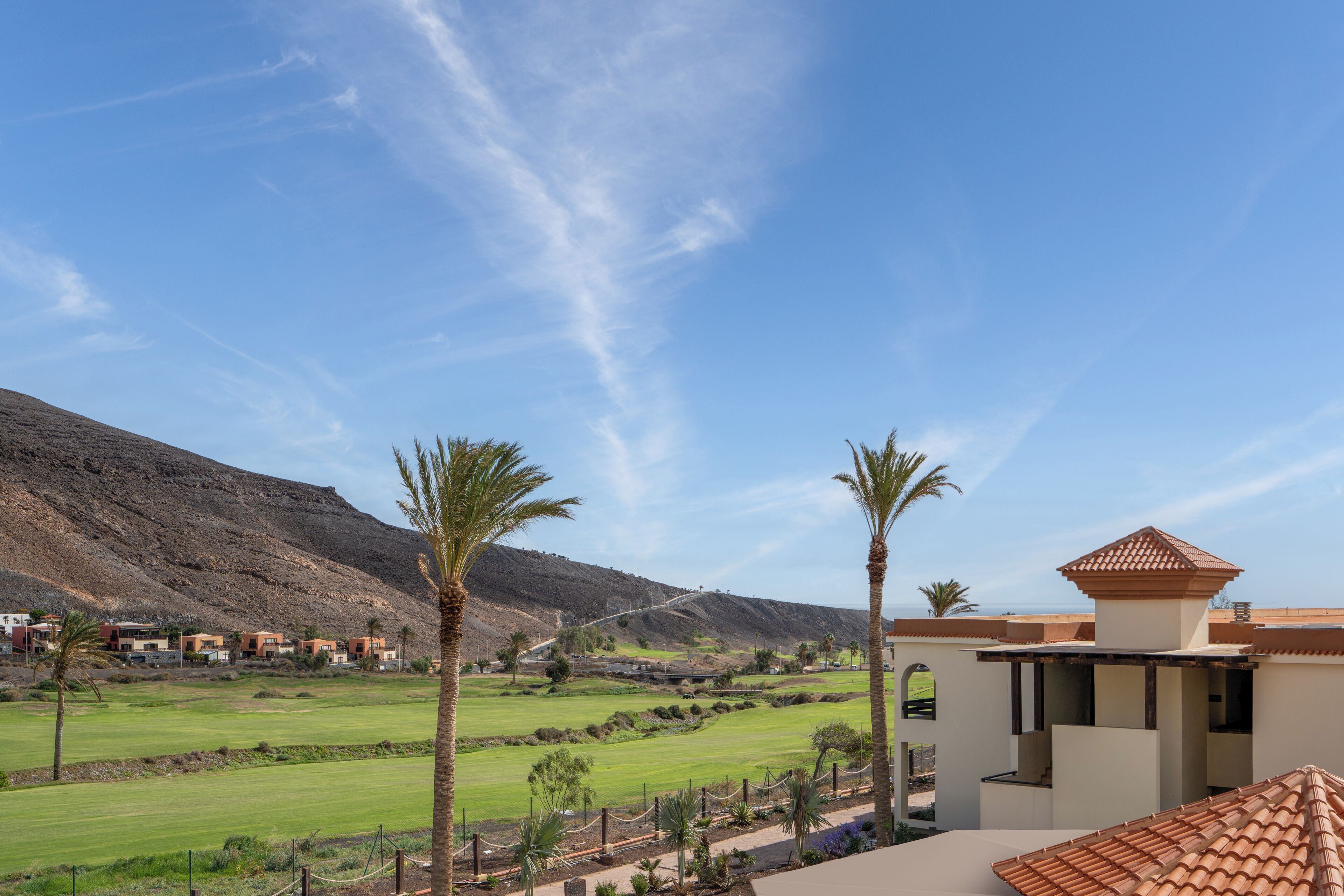 A view of a golf course with a mountainous backdrop, palm trees, and a building with a tiled roof.