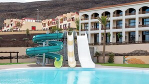 Una piscina con toboganes, un edificio con balcones y montañas al fondo.