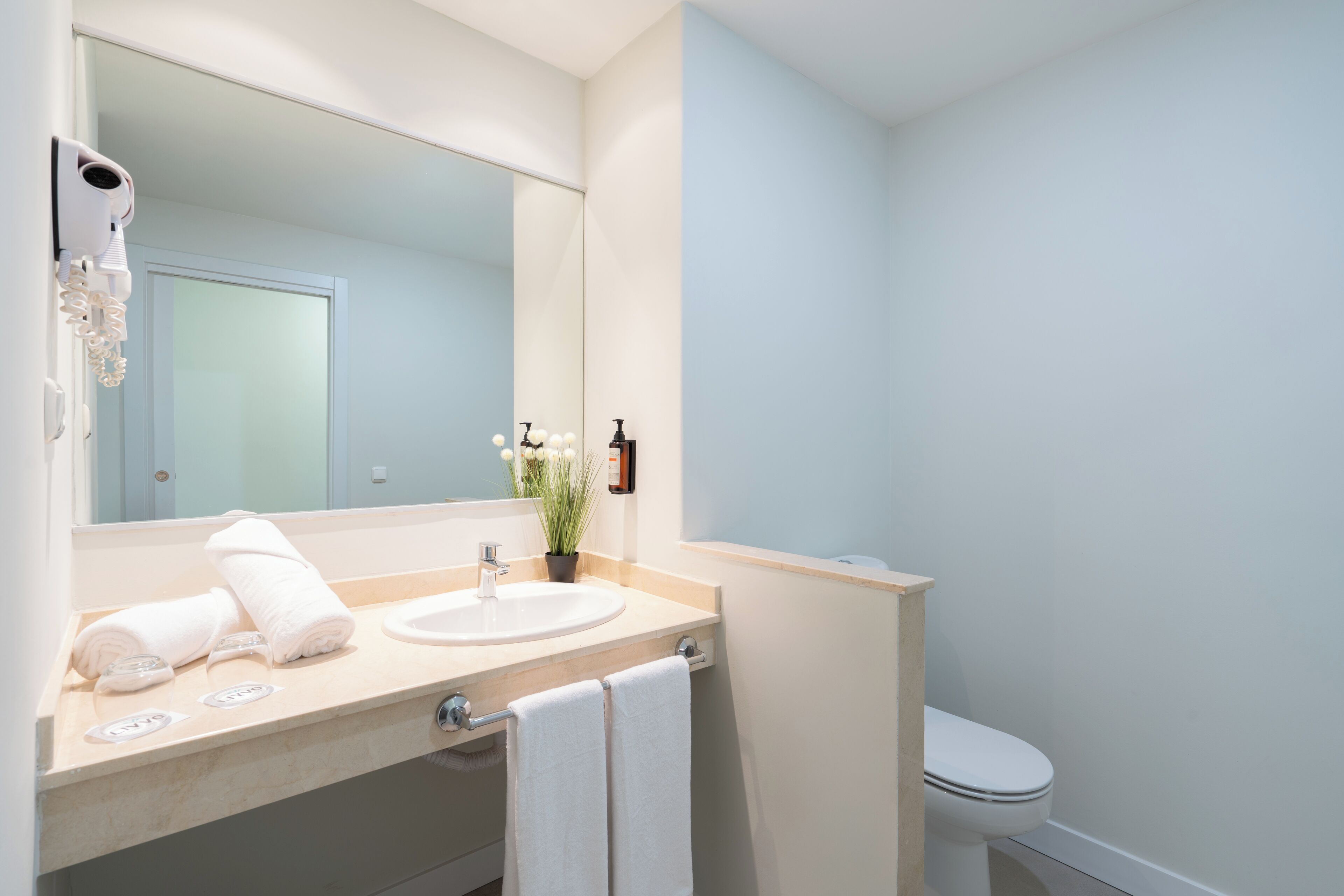 Une salle de bain moderne avec un grand miroir, un lavabo, des toilettes et un sèche-cheveux fixé au mur.