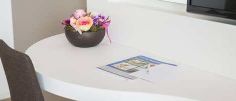 Une table blanche avec un bol noir contenant des fleurs, une brochure et une chaise.