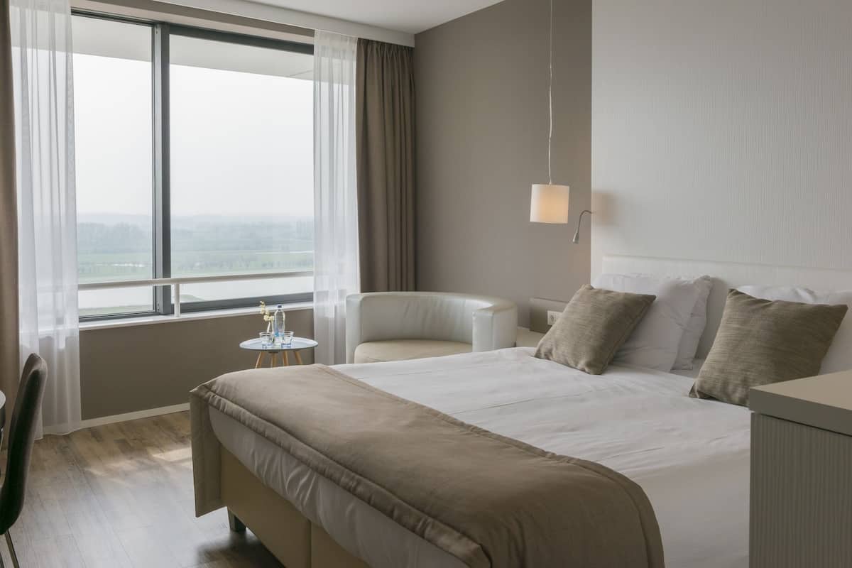 Quarto de hotel com uma cama grande, uma cadeira, uma mesinha com copos e vista para o exterior pela janela.