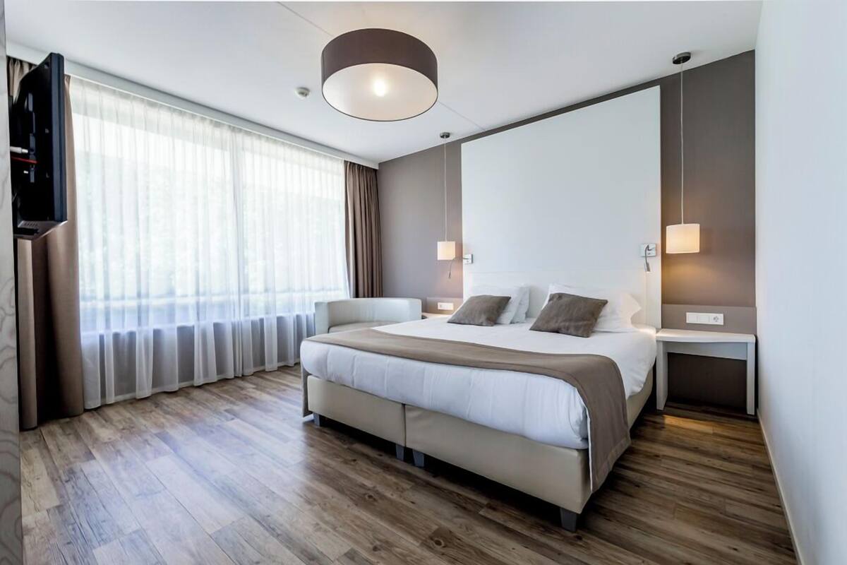 Quarto de hotel moderno com uma cama grande, televisão de tela plana, mesa de cabeceira e uma janela ampla com cortinas.